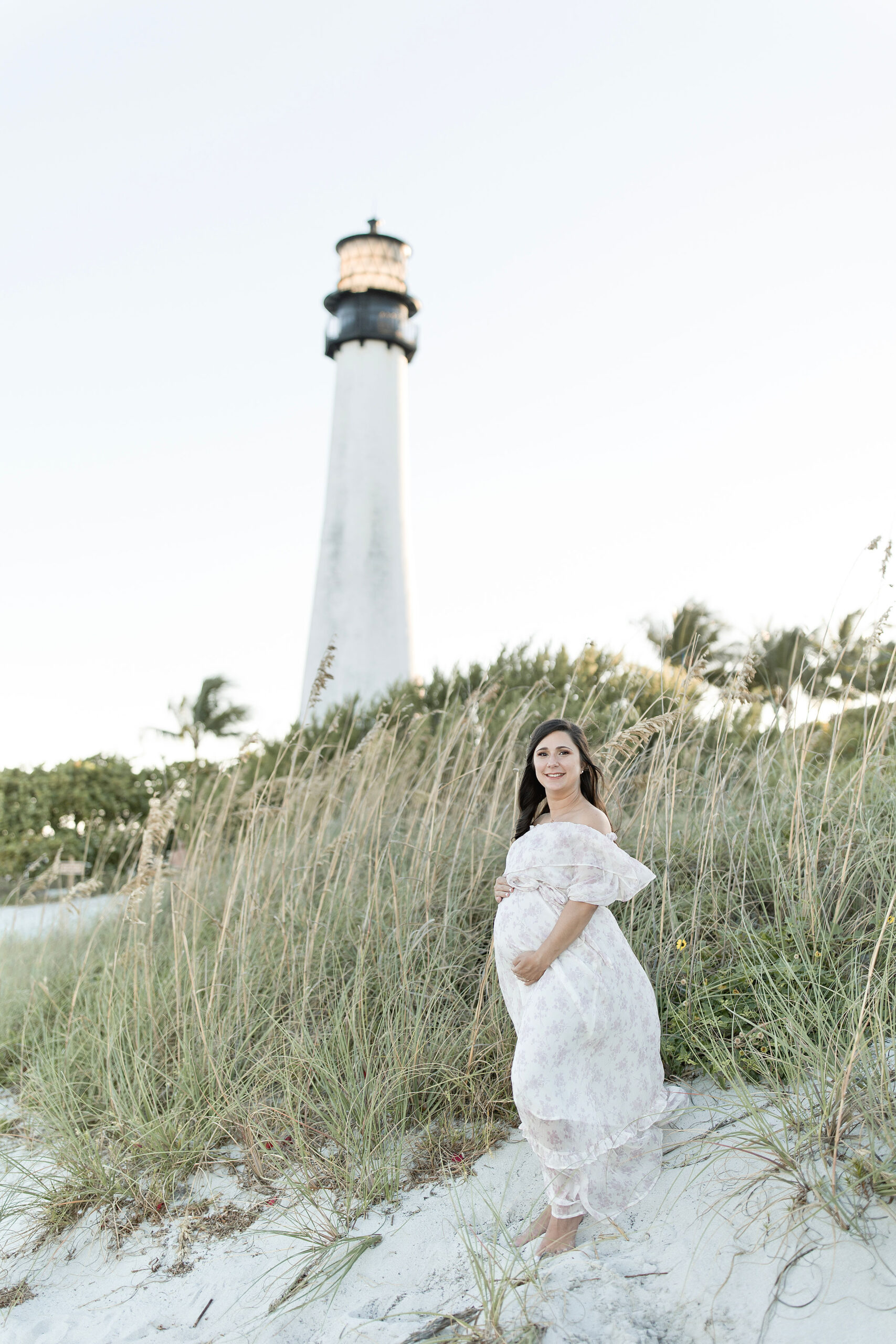 A mom to be in a floral maternity dress stands on a windy beach dune in front of a lighthouse after some miami prenatal yoga