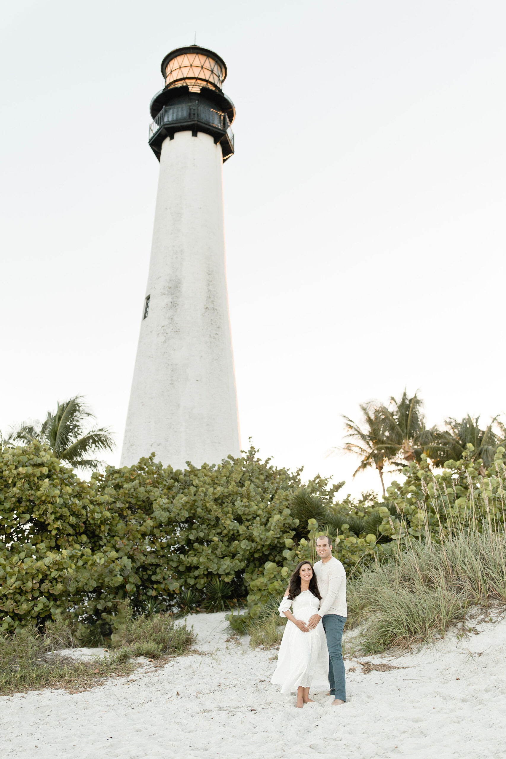 A mom to be in a white dress stands on a beach by the dunes under a lighthouse with her husband after some miami prenatal yoga