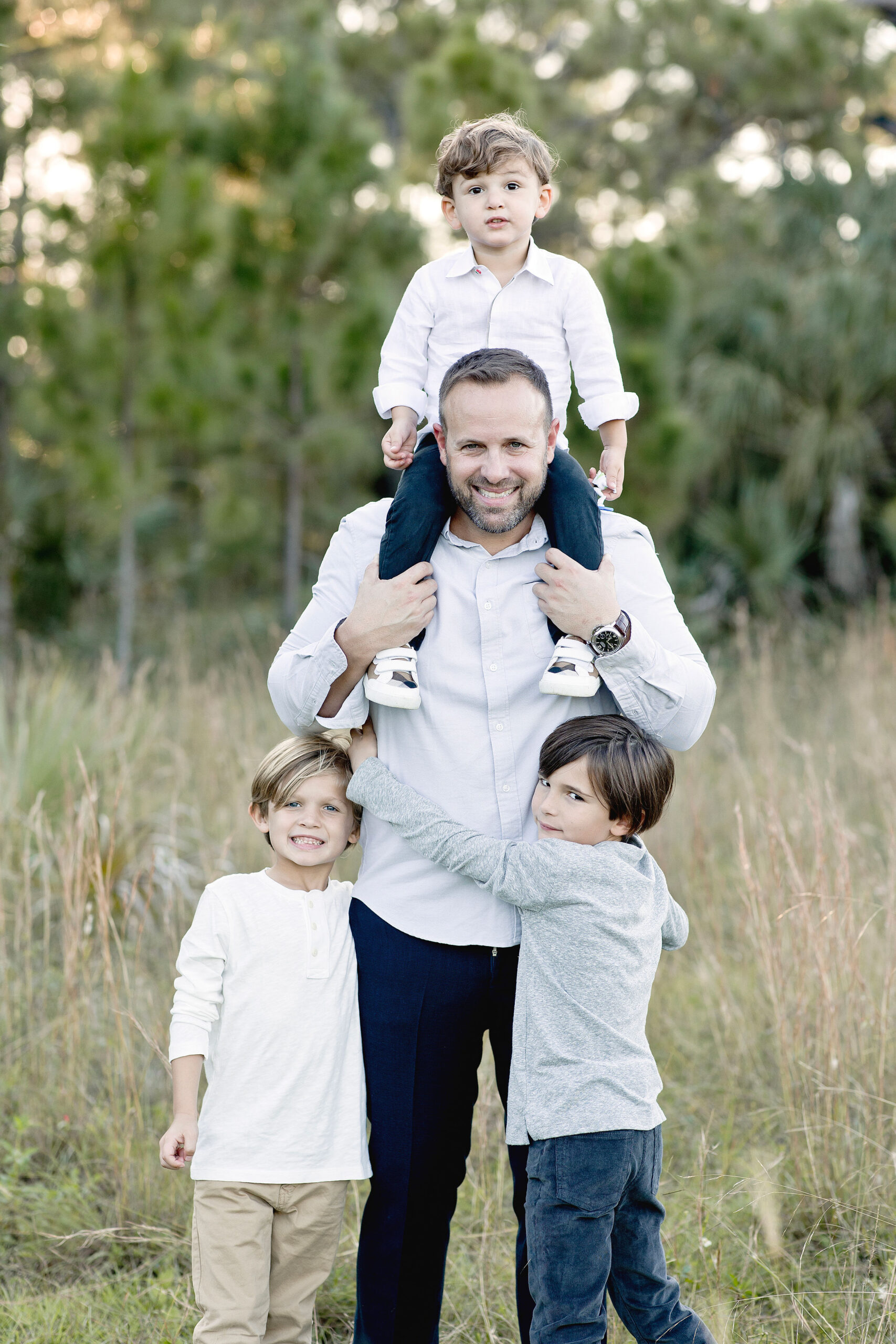 A father stands in a field with his toddler son on his shoulders and two older sons hugging him