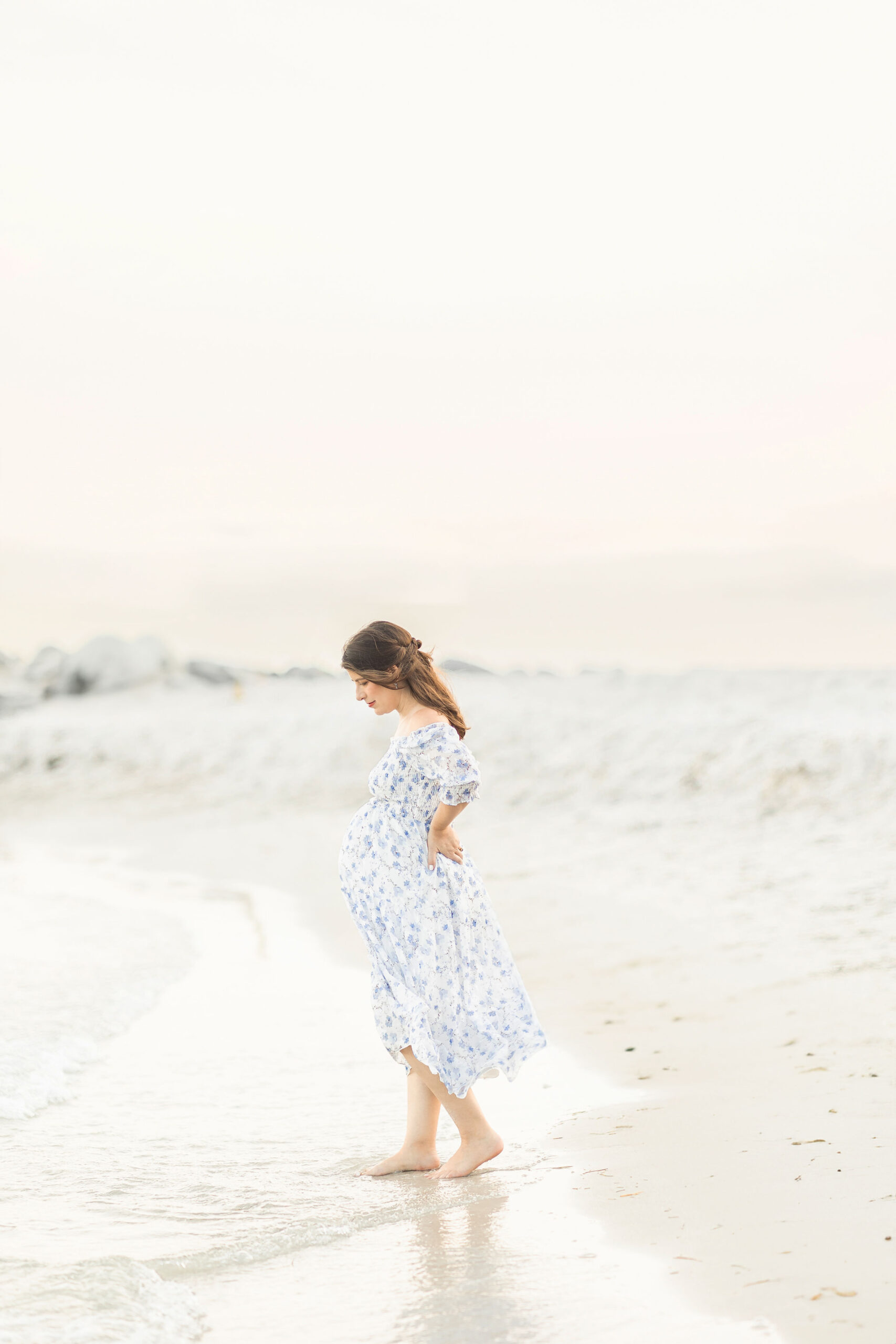 A mom to be walks in the water on a beach in a blue and white maternity dress