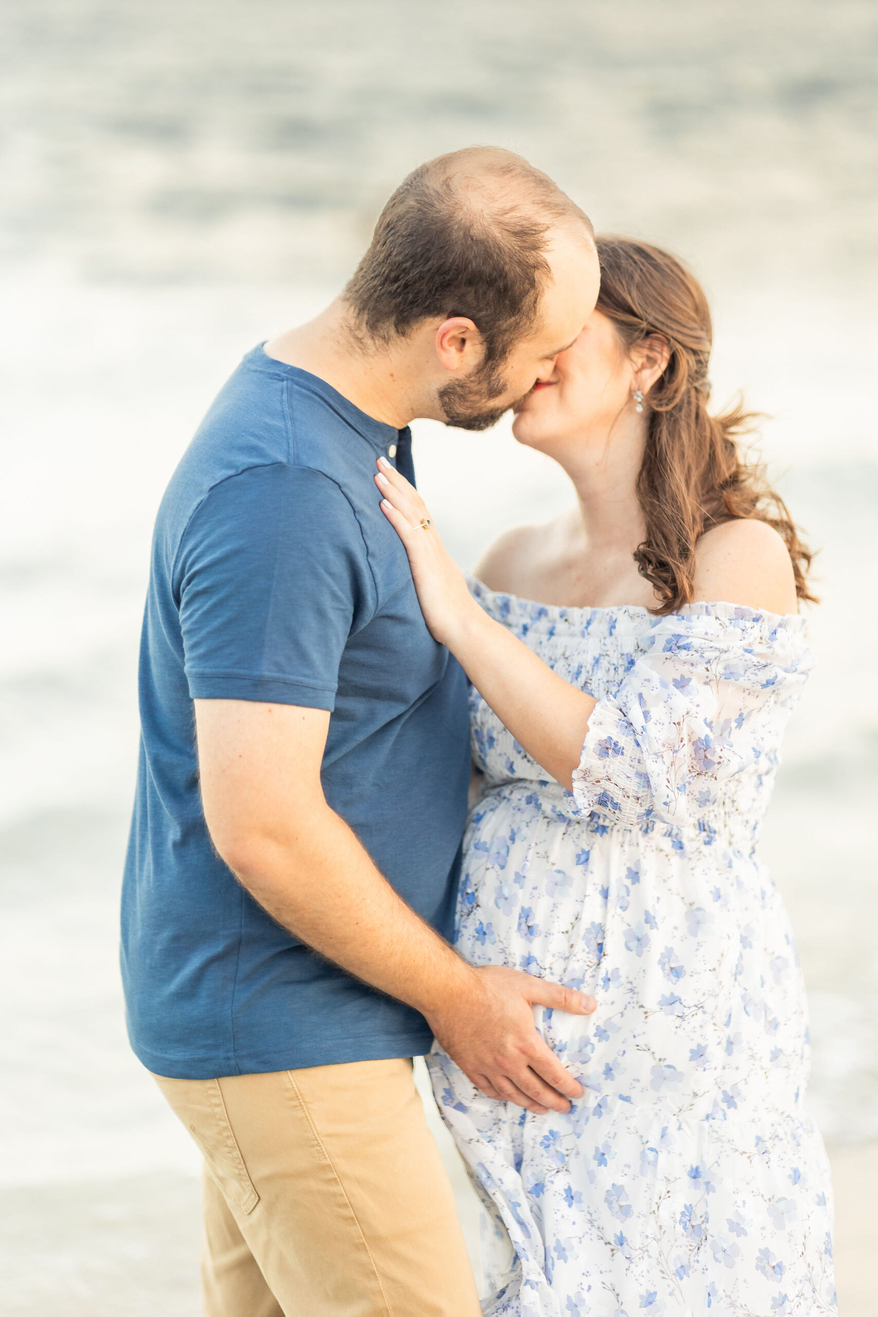 Expecting parents stand on a beach sharing a kiss while holding the bump after visiting coastal midwifery