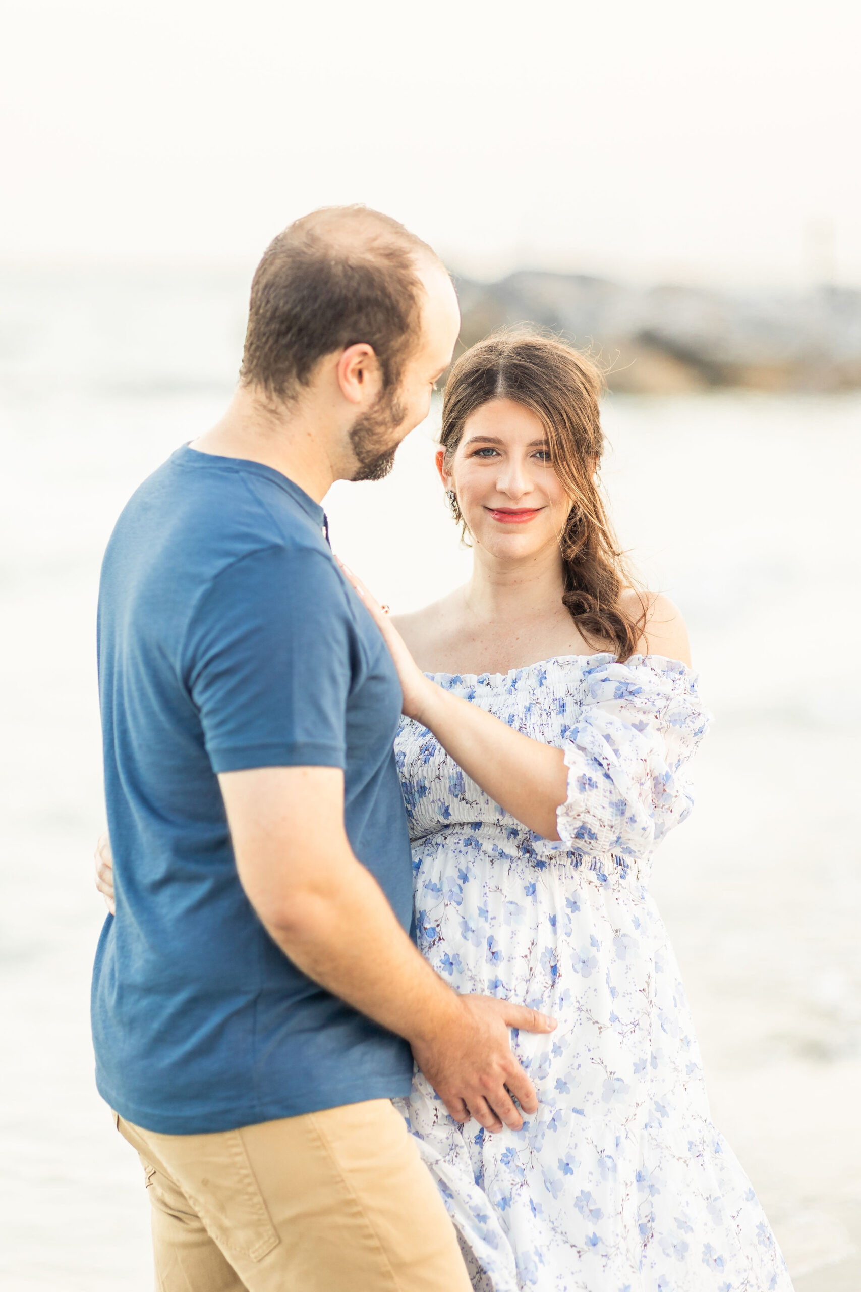 A mom to be leans onto her husband in a blue dress while standing on a beach at sunset before visiting coastal midwifery