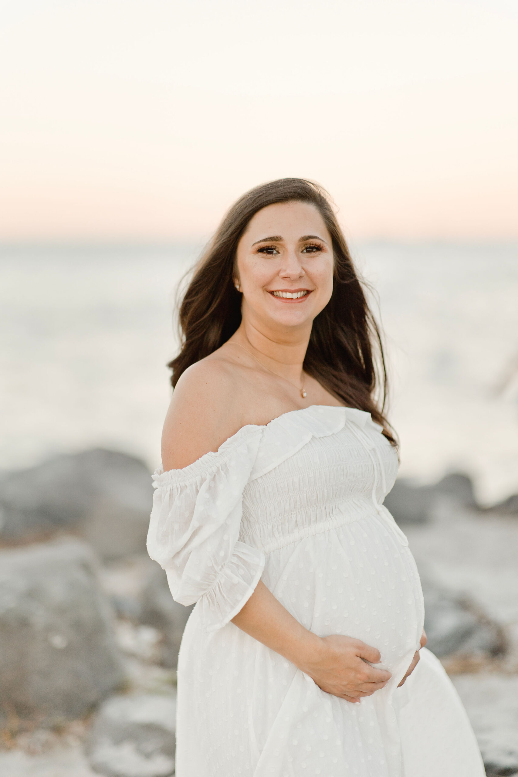A mom to be in a white maternity gown stands on a rocky beach holding her bump before visiting east coast midwifery
