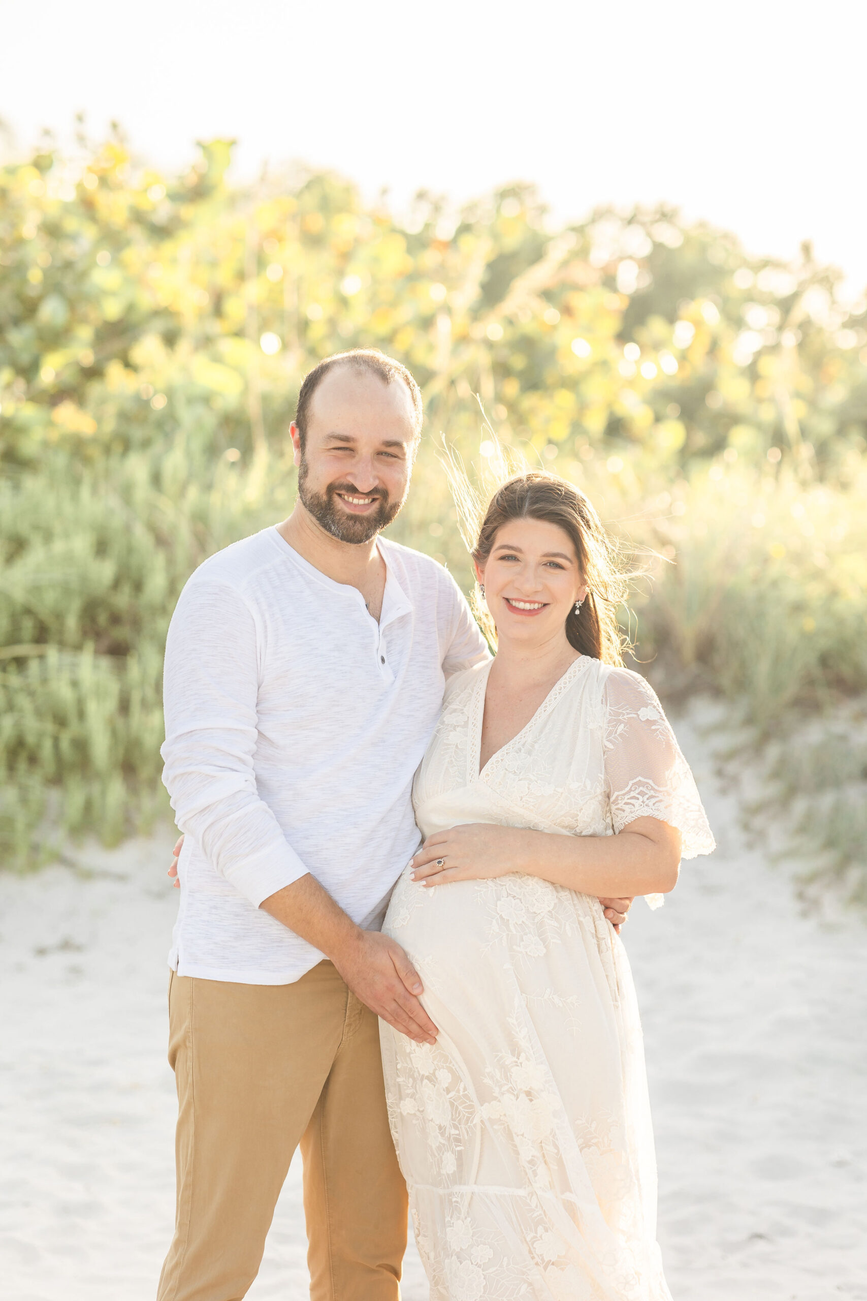 A mom to be stands in a white lace dress on a beach with her husband holding the bump at sunset before visiting south miami hospital maternity