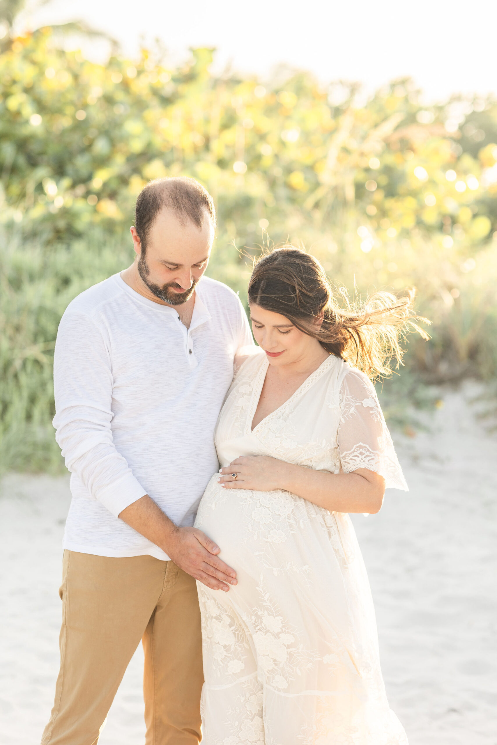 Expecting parents smile down at the bump while standing on a beach at sunset after visiting south miami hospital maternity