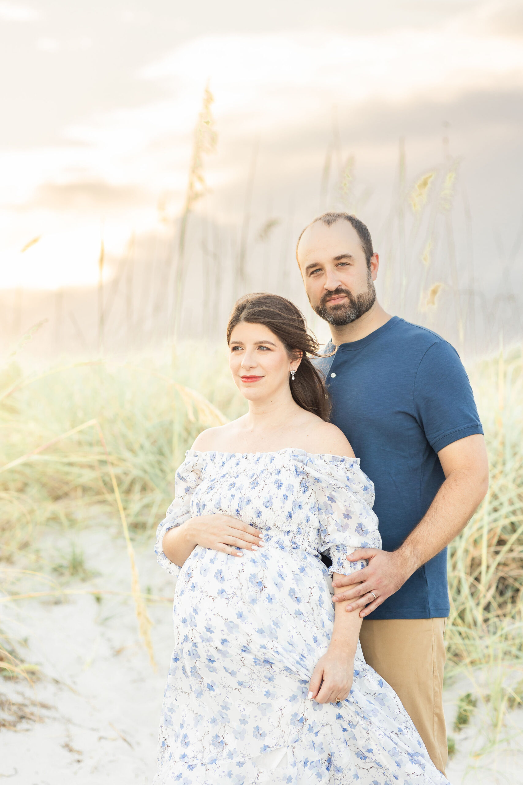 An expecting father in a blue shirt stands behind his pregnant wife on a beach at sunset