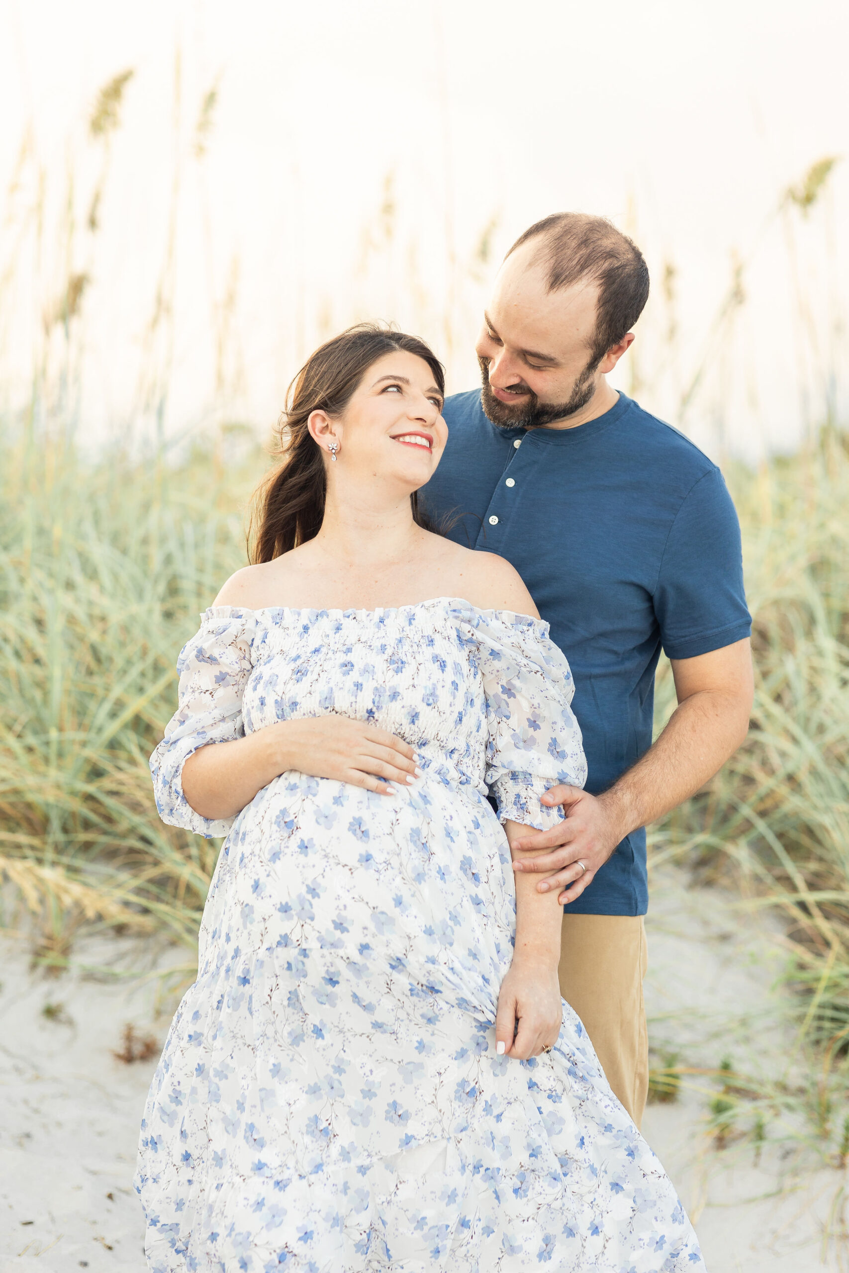Expecting parents stand on a beach at sunset smiling at each other and holding the bump after meeting amazing births and beyond