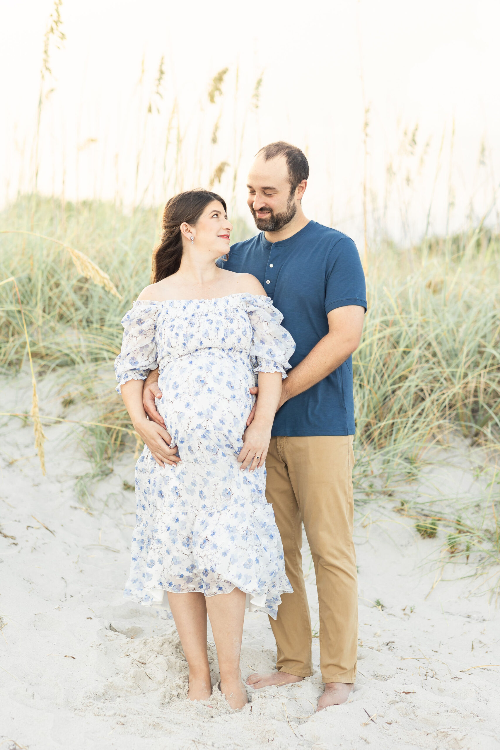 A mother to be in a blue floral print maternity dress stands on a beach smiling up to her husband holding the bump after amazing births and beyond