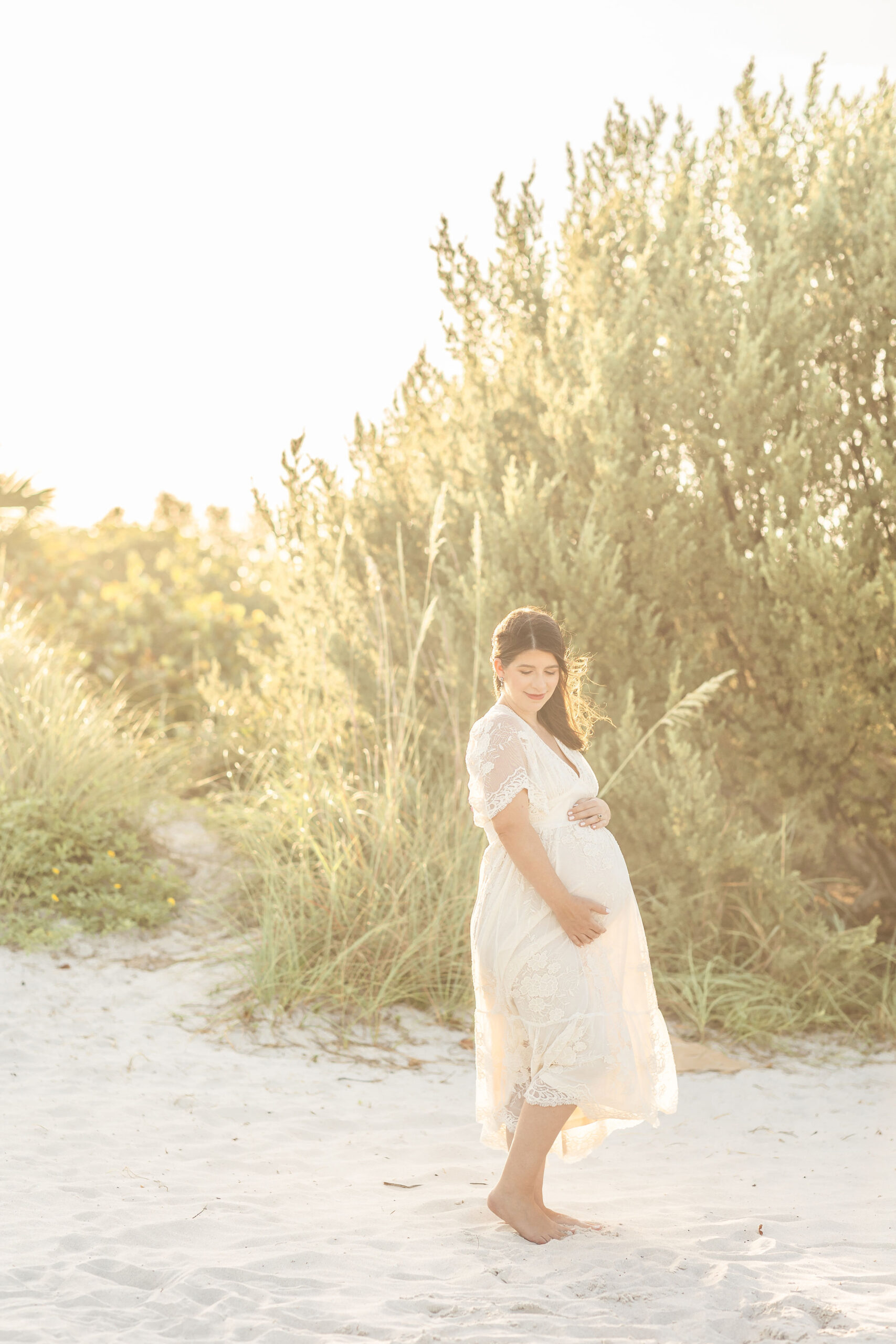 A mother to be walks down a windy beach at sunset by the dunes in a white maternity gown