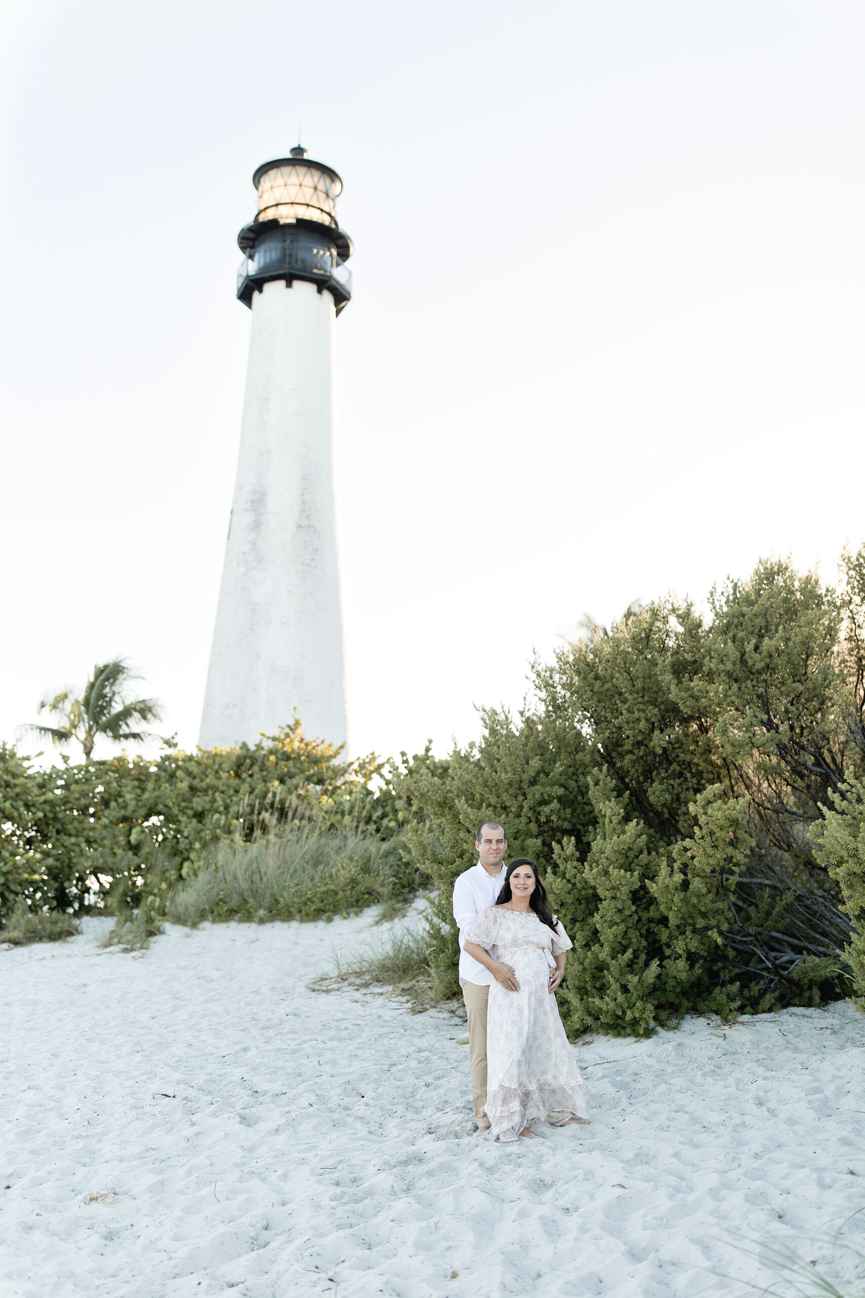 A mother to be and her husband stand on a beach dune at sunset holding the bump in front of a lighthouse after visiting natural birthworks birth center
