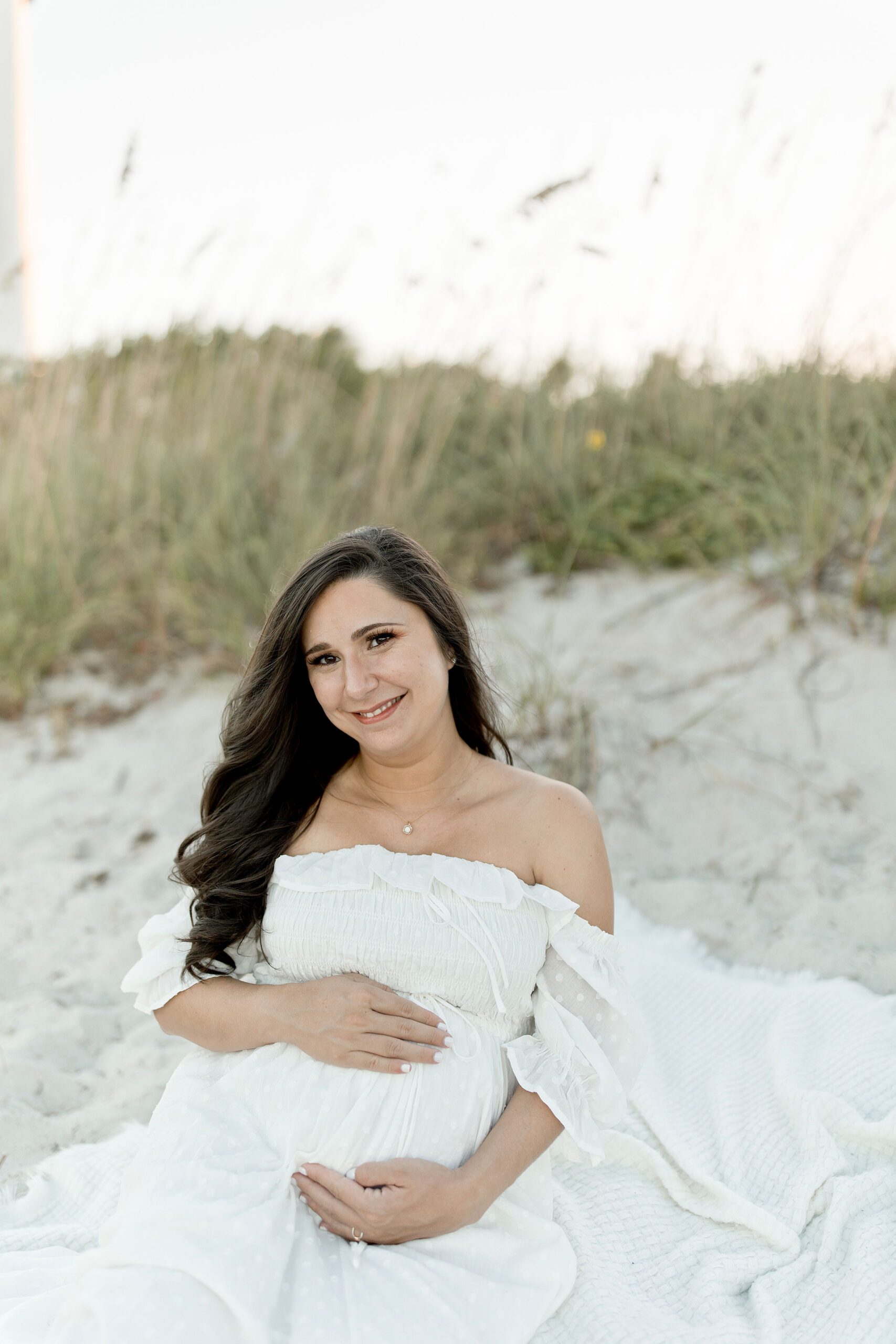 A mother to be in a white maternity gown sits on a beach blanket in the dunes holding her bump after visiting natural birthworks birth center