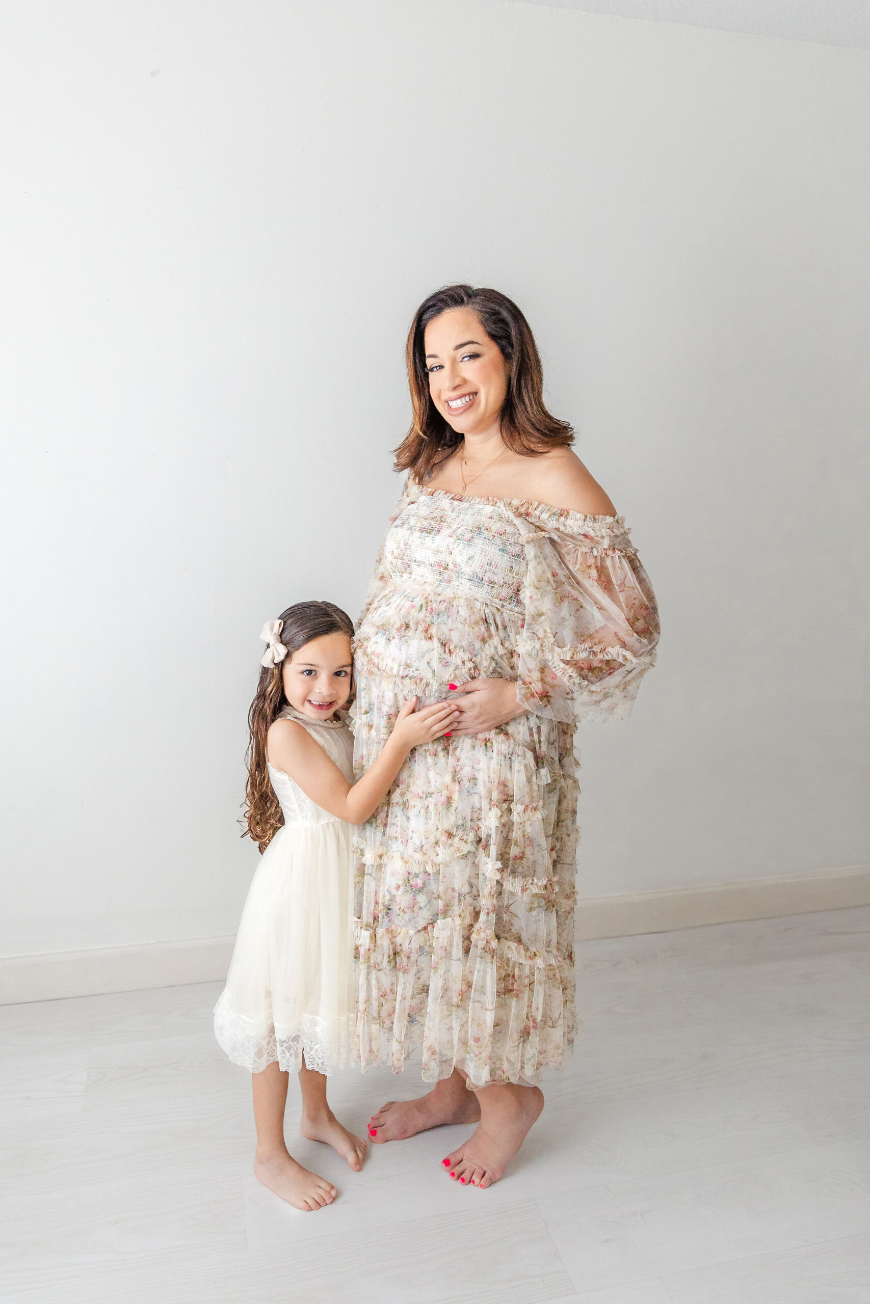 A pregnant mother stands in a studio with her toddler daughter in a white dress as they hug the bump