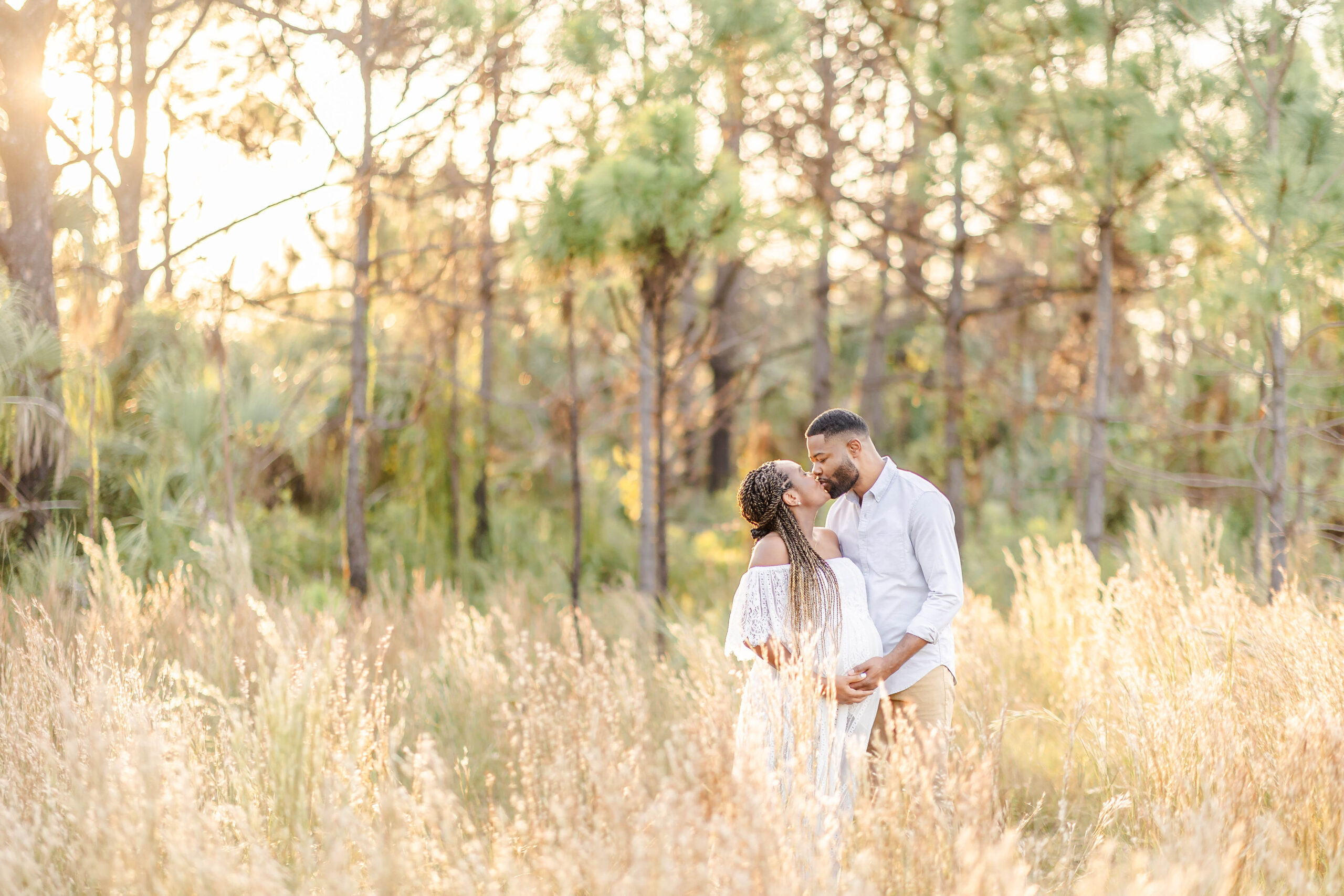 Expecting parents hold the bump while standing in a field of tall golden grass at sunset and kissing after meeting eyla cuenca