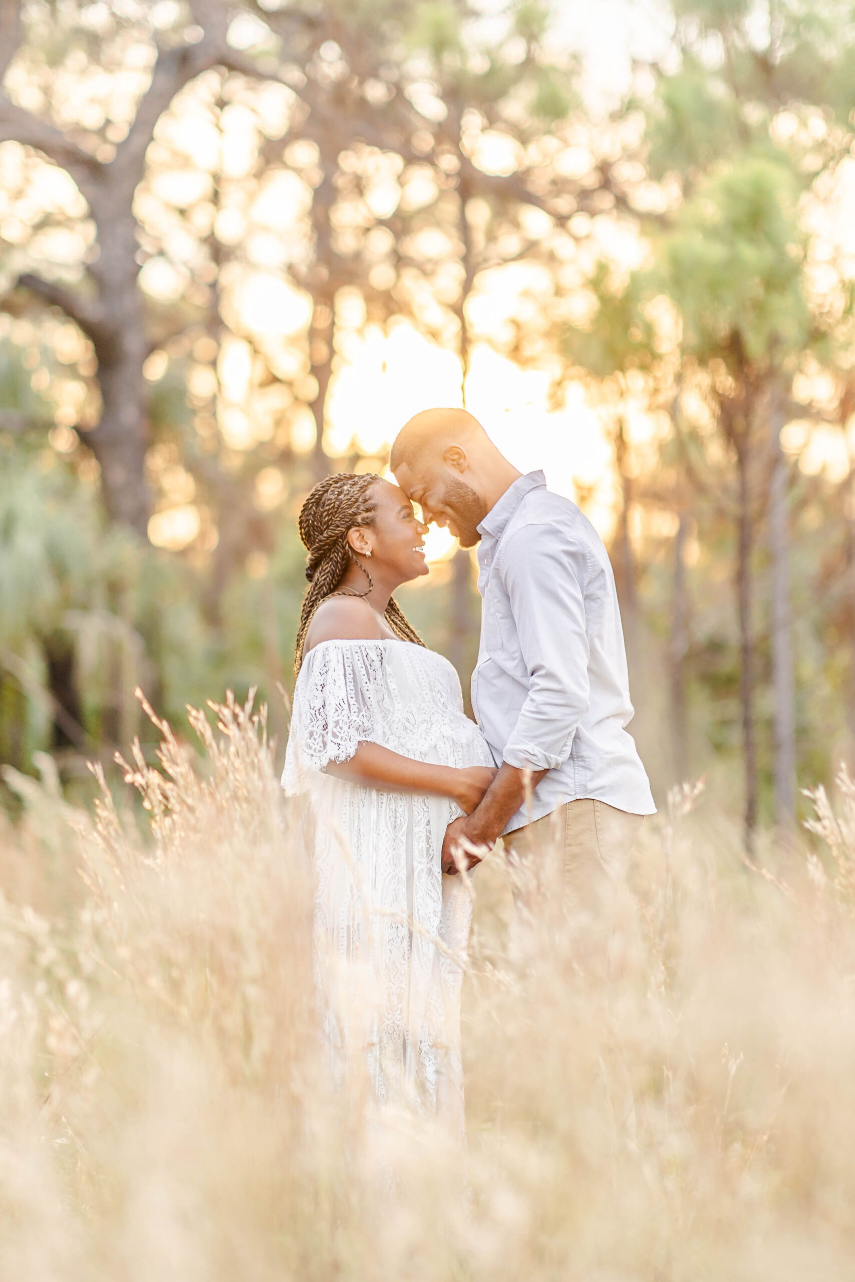 Happy expecting parents stand in a field of tall golden grass at sunset holding the bump before meeting eyla cuenca