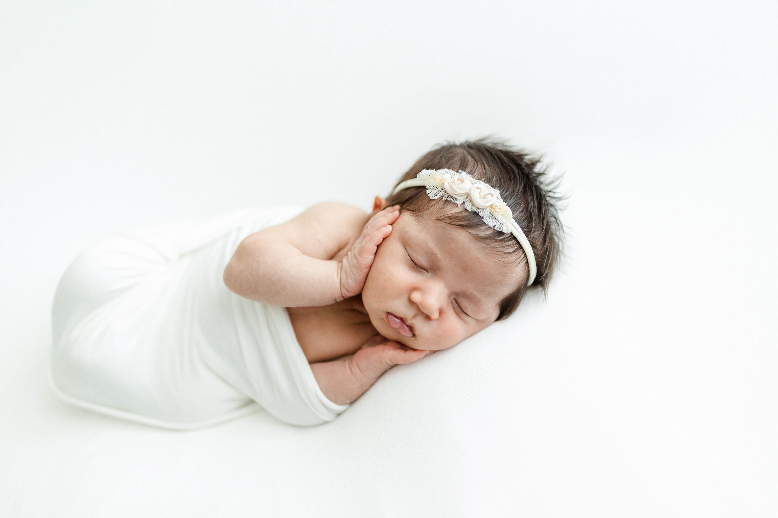 A newborn baby sleeps in a white swaddle with hands on her face