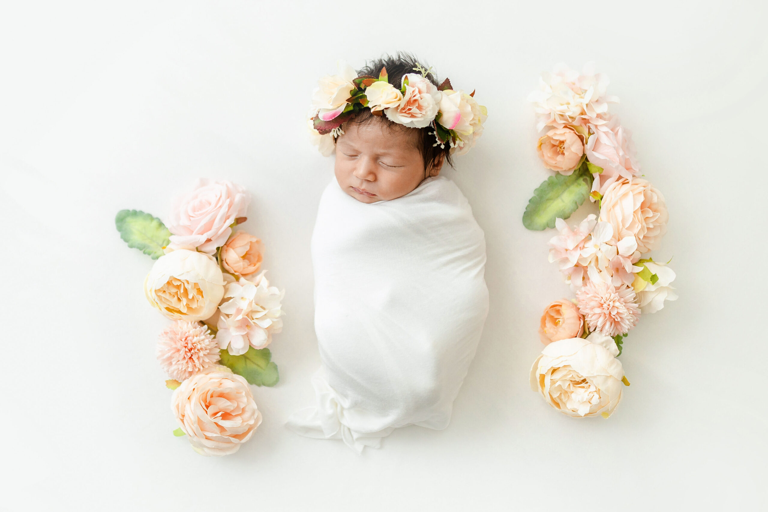 A newborn baby sleeps in a white swaddle and floral headband surrounded by matching flowers in a studio thanks to mom's breast friend