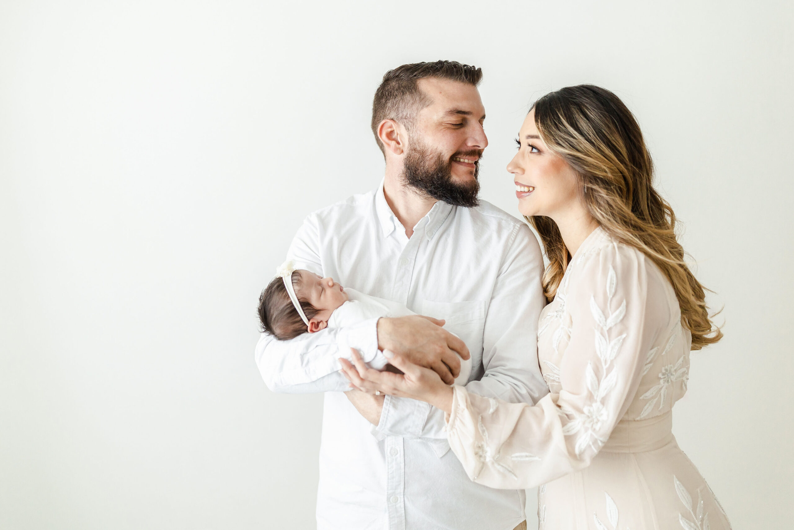 Happy new parents smile at each other while holding their sleeping newborn baby in a studio after meeting mom's breast friend