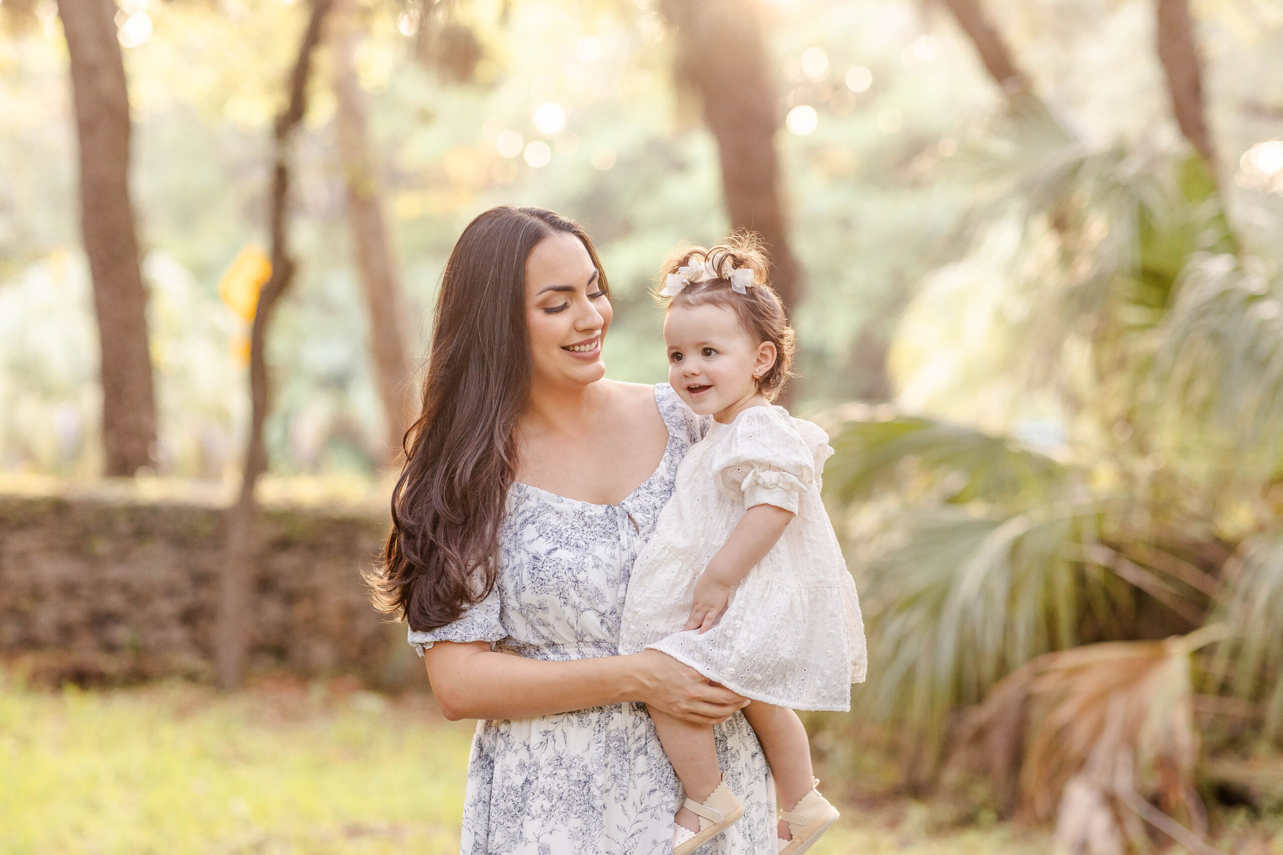 A mother in a floral print dress stands in a park at sunset holding her happy toddler daughter in a white dress