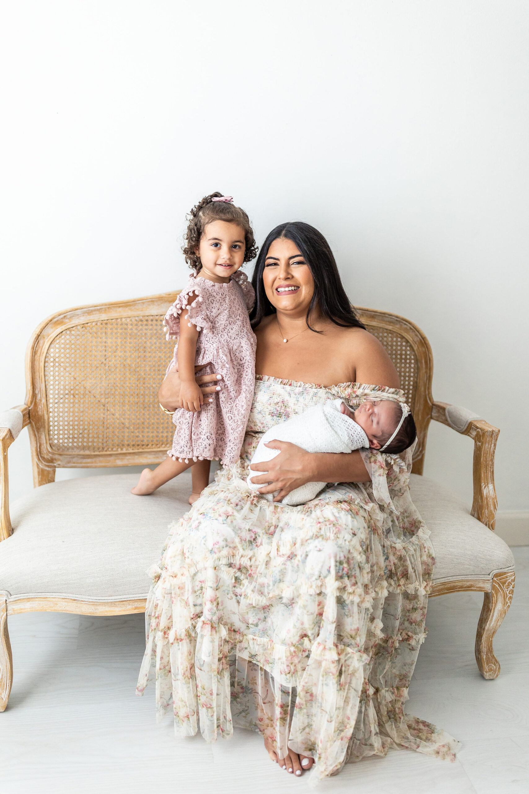 A happy mother in a long floral dress sits on a bench in a studio with her toddler daughter in a pink dress and newborn baby sleeping in one arm
