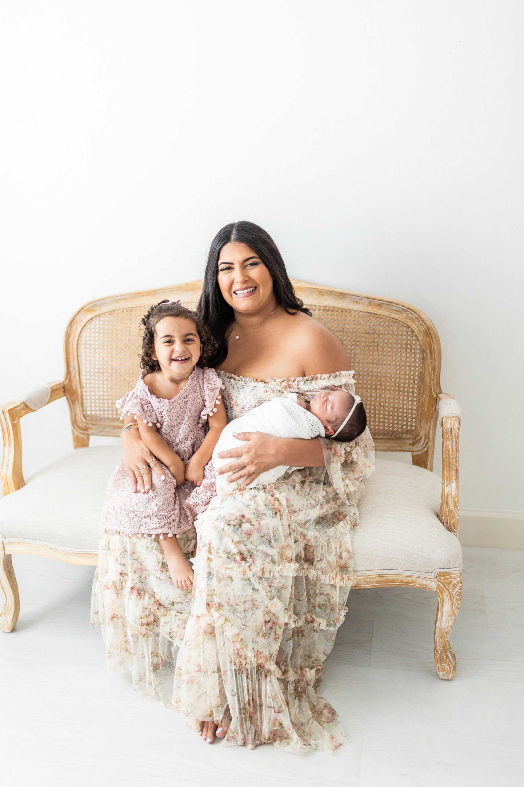 A happy mom sits on a bench in a studio with her toddler daughter on one leg and her newborn baby sleeping in her other arm after meeting mamalactea