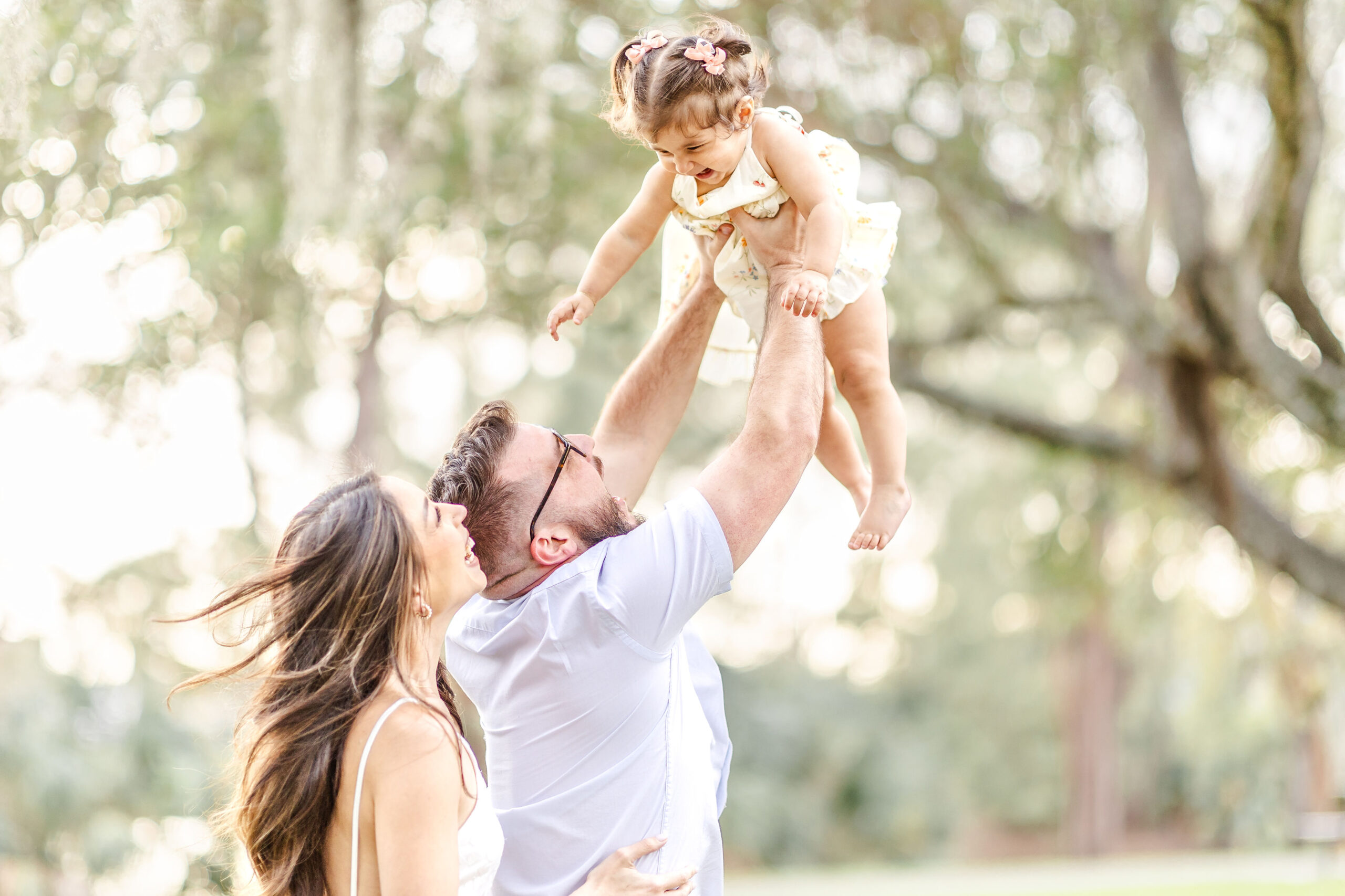 A happy dad laughs with his toddler daughter as he lifts her above his head with mom