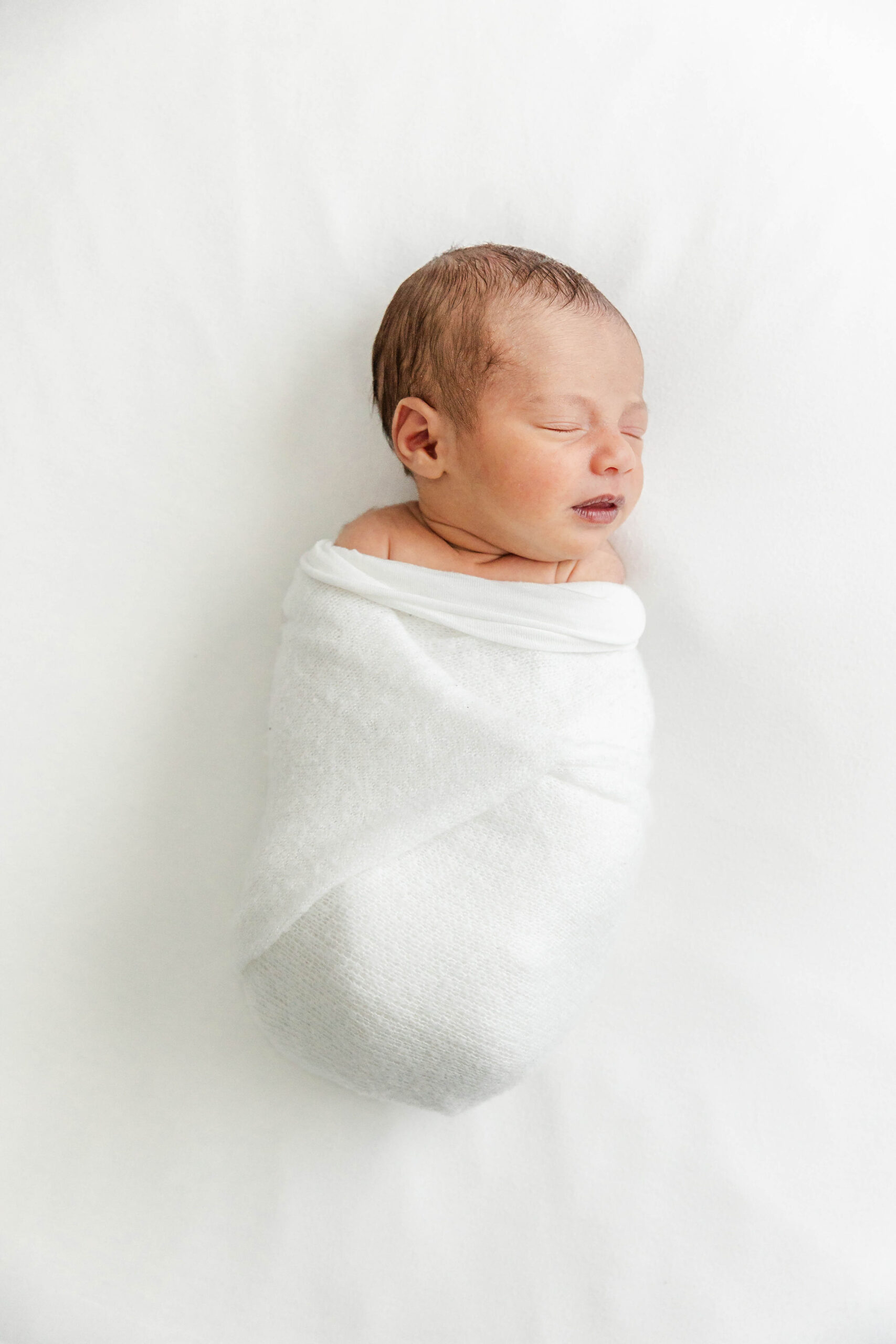 A newborn baby sleeps in a white swaddle on a bed in a studio