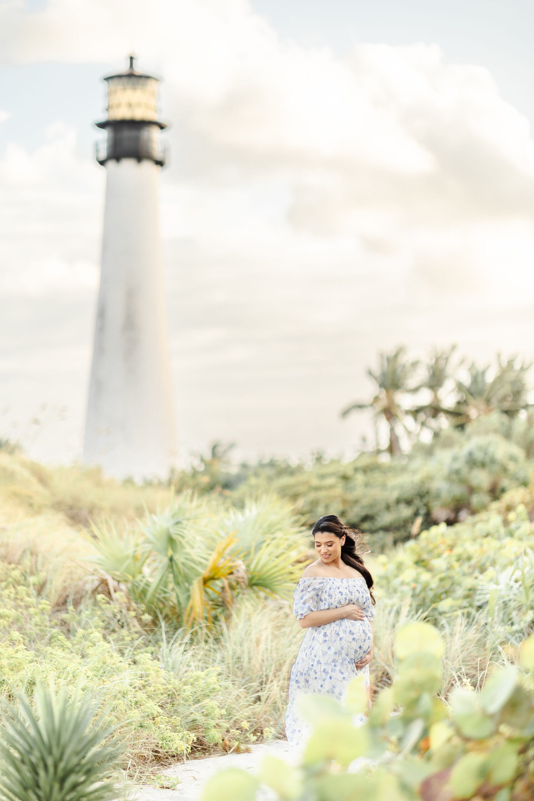 A mother to be walks in a windy beach path by a lighthouse while holding her bump at sunset thanks to a fertility clinic miami