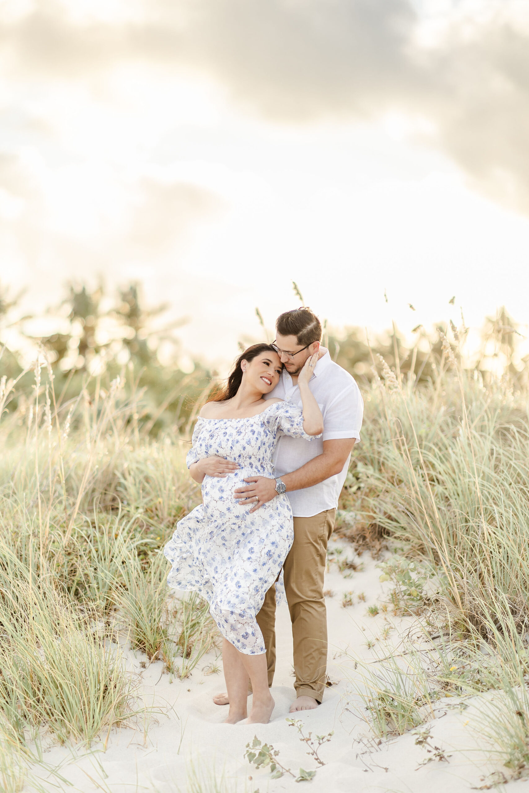 A mom to be leans into the chest of her husband while standing in a beach dune at sunset thanks to a fertility clinic miami