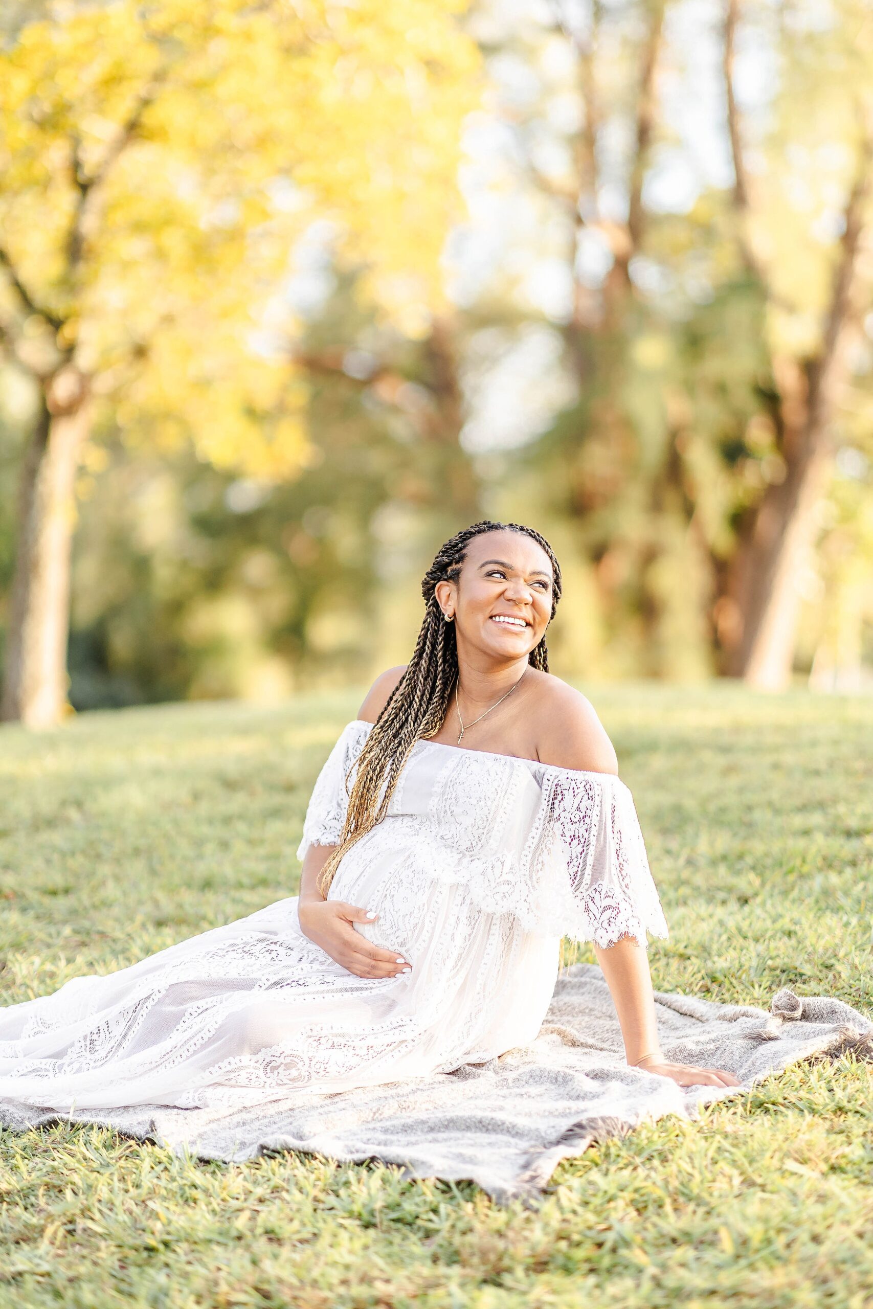A happy mother to be in a white lace dress sits on a picnic blanket in a park at sunset after visiting a miami obgyn