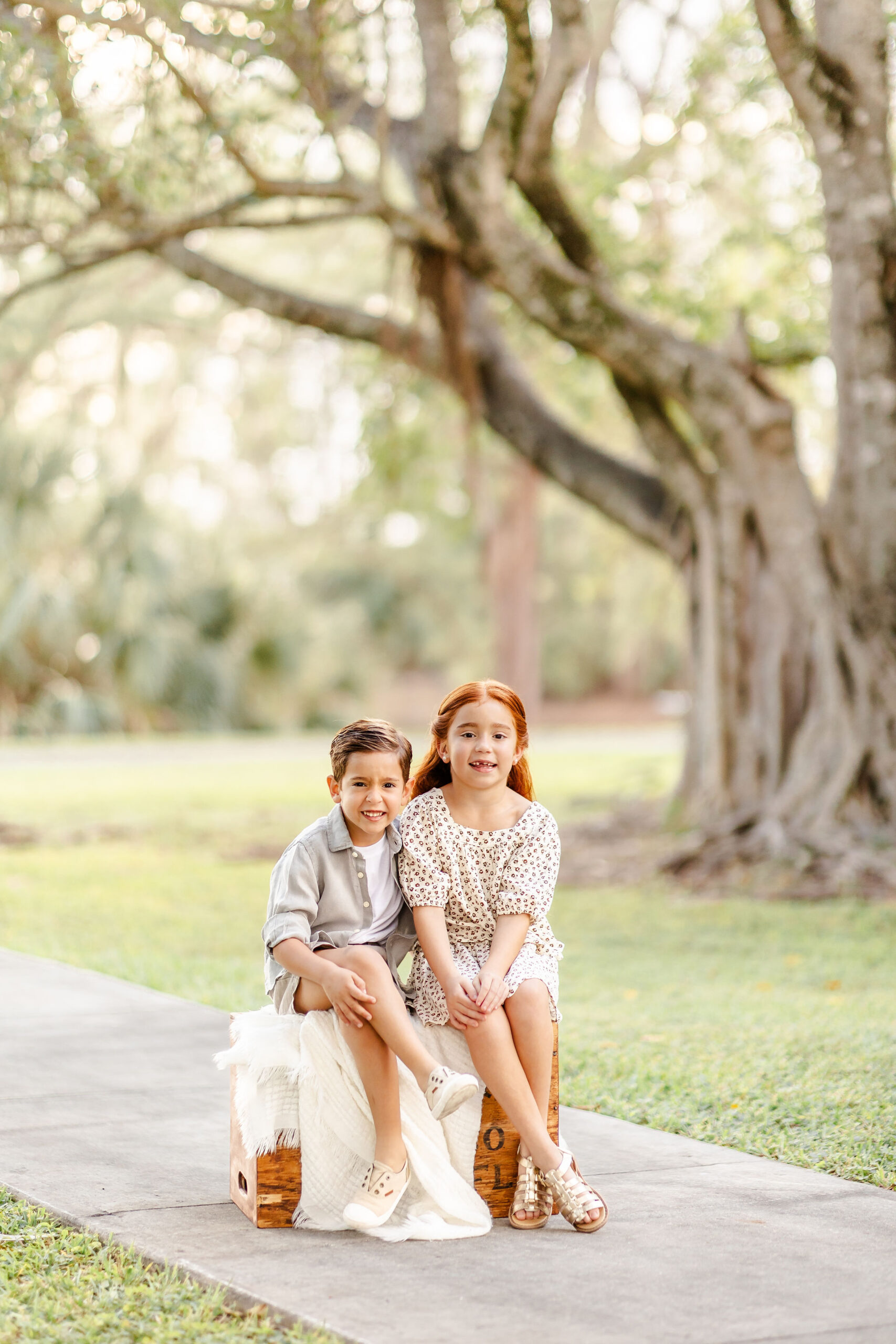 A young boy sits on a wooden box with his sister in a park sidewalk at sunset