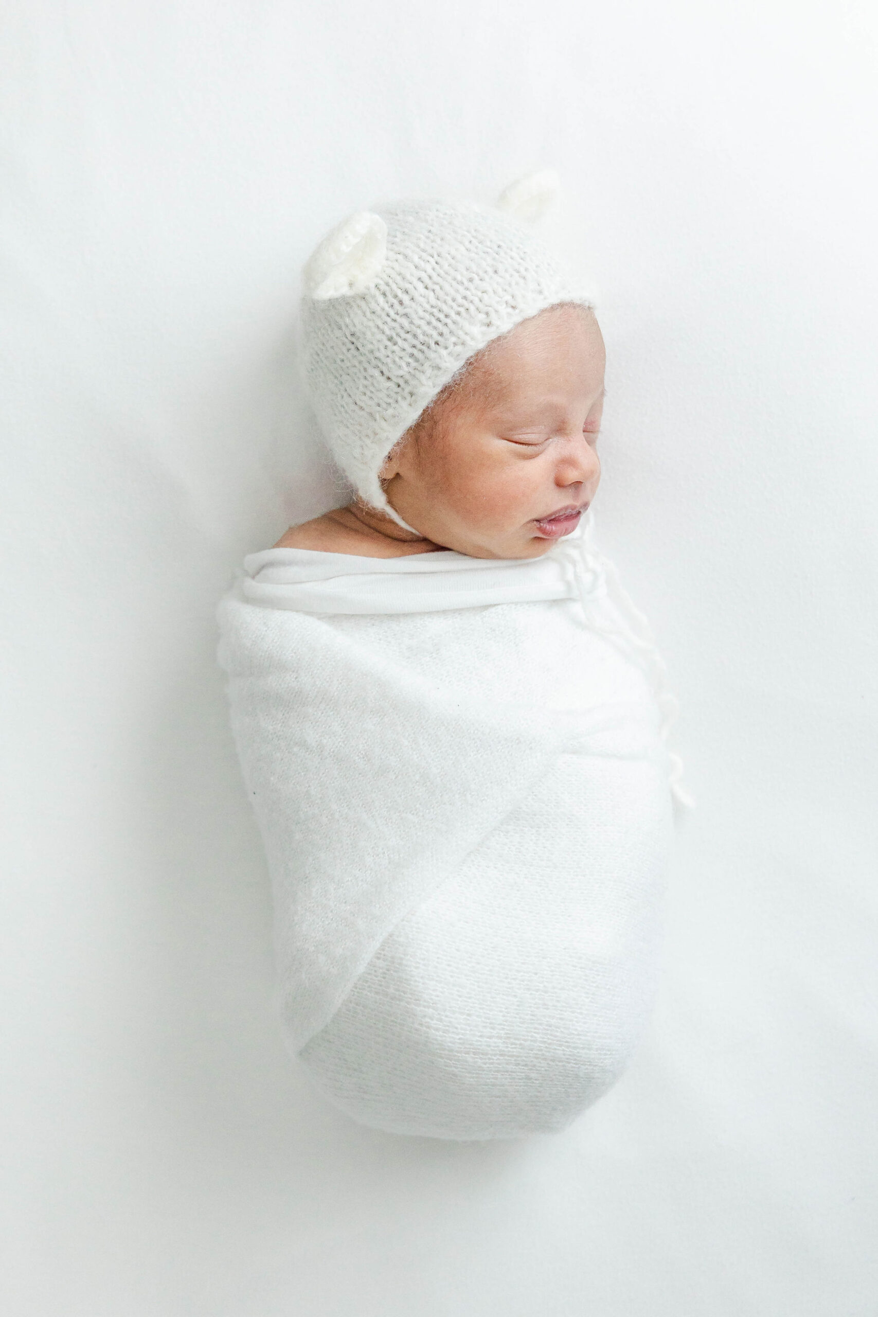 A newborn baby sleeps in a white bonnet with ears on a white bed in a studio after visiting a pediatric chiropractor miami