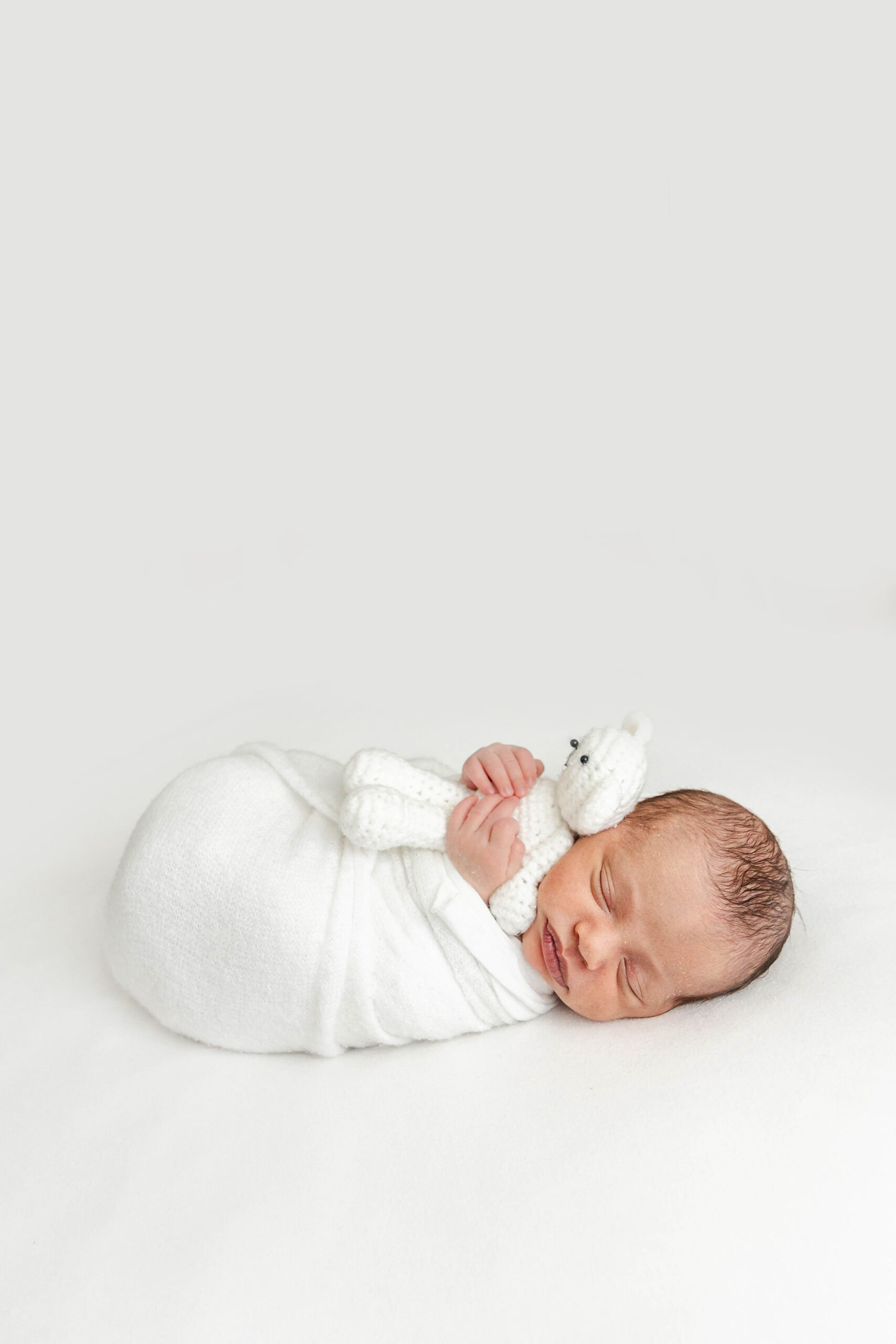 A newborn baby sleeps while clutching a white knit bear in a white swaddle thanks to a pediatric chiropractor miami