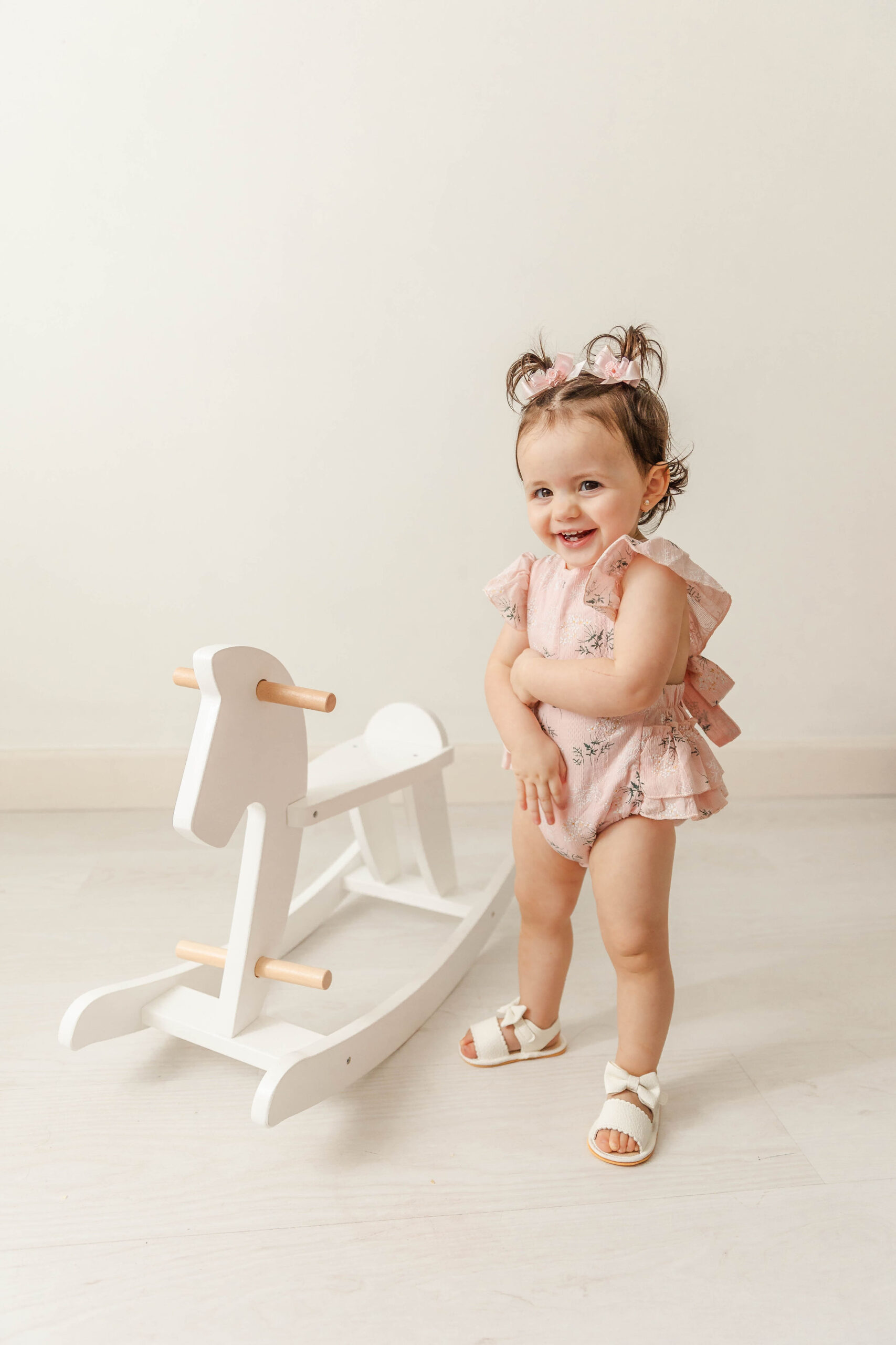 A happy toddler girl in a pink onesie stands in a studio with a white wooden rocking horse