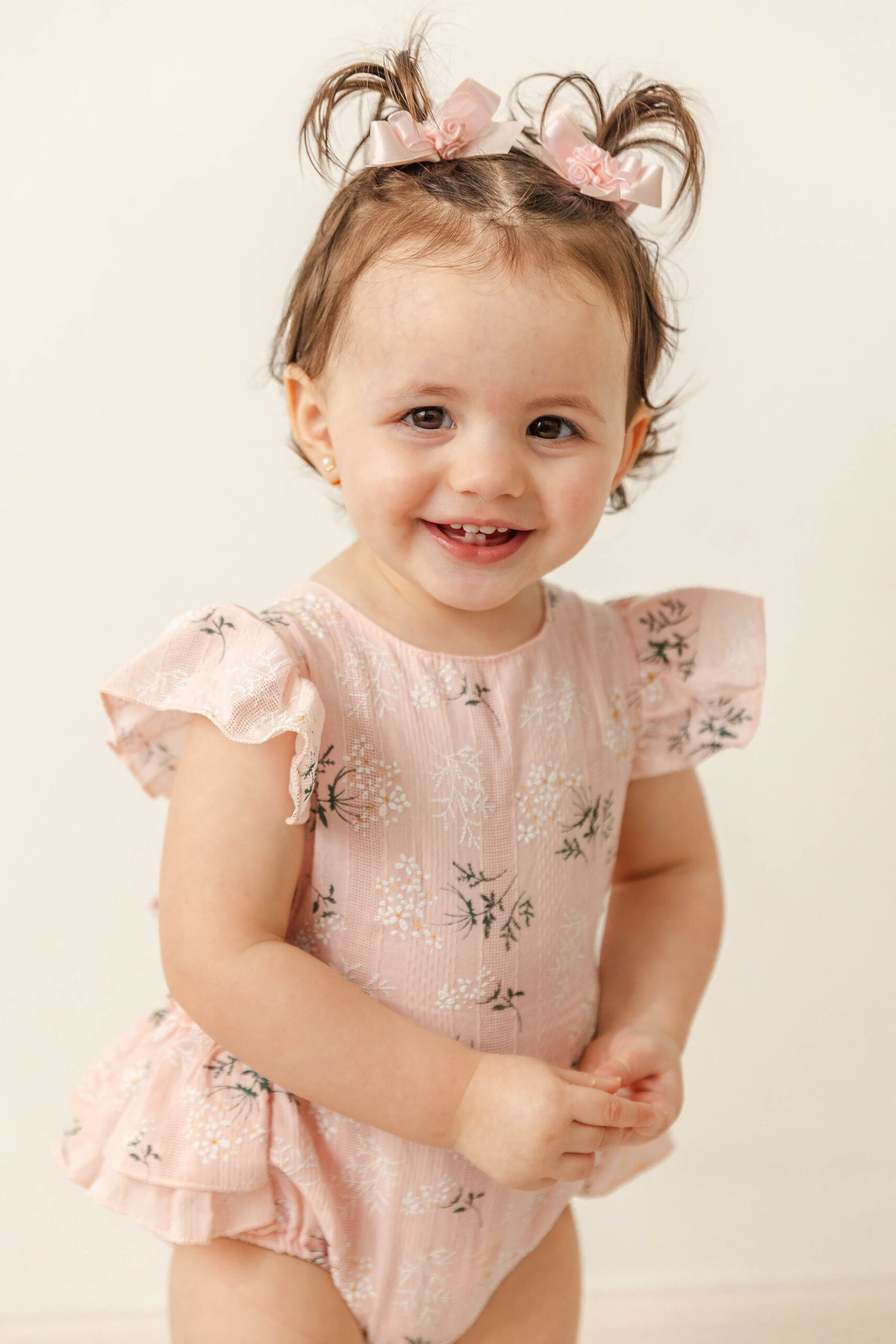 A giggling toddler girl with tiny pig-tails stands in a studio in a pink onesie before some baby music classes miami