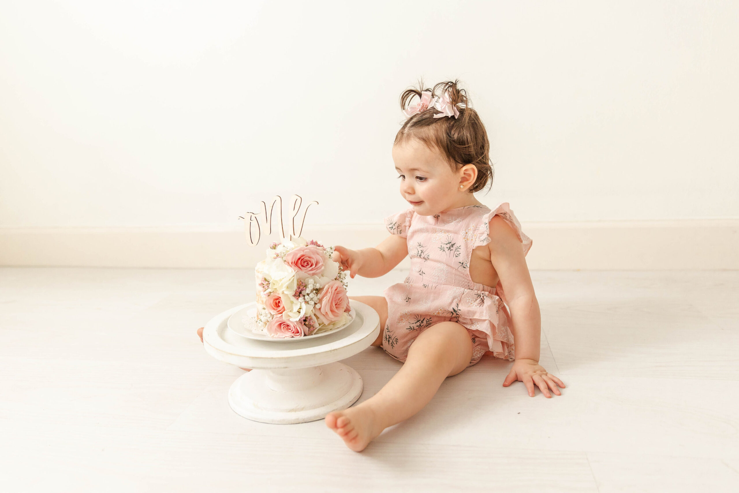 A toddler girl sits on the floor while examining a flower covered cake in a pink onesie for her birthday before some baby music classes miami