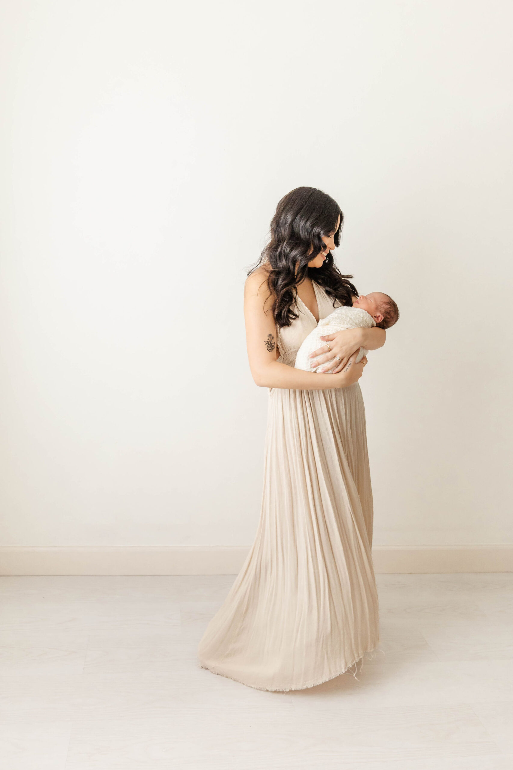 A smiling mother in a beige gown walks in a studio while holding her sleeping newborn baby