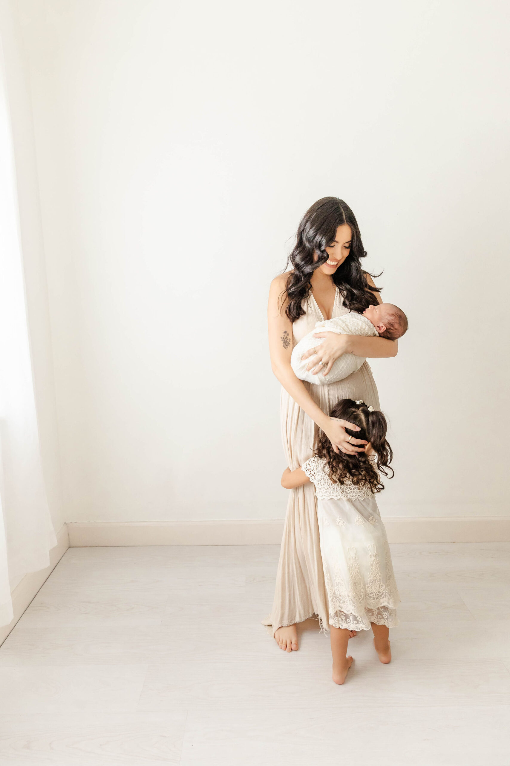 A toddler girl hugs her mom while standing in a studio holding her newborn baby thanks to pelvic floor therapy miami