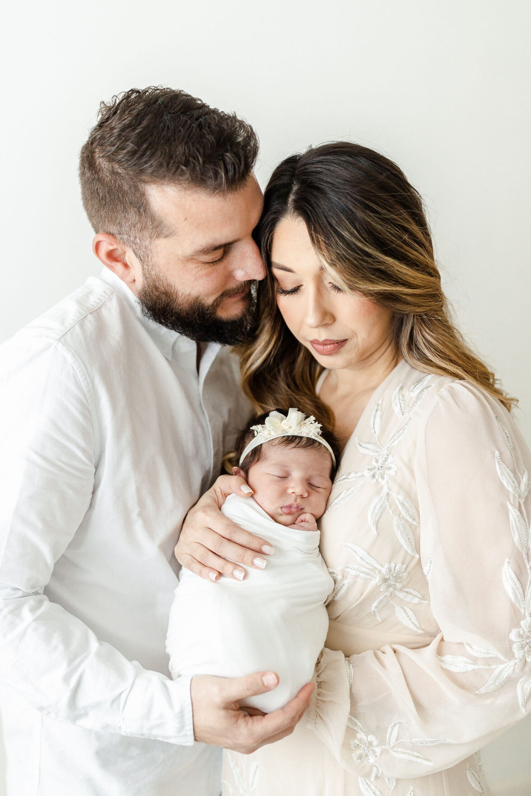 Happy new parents in white cradle their sleeping newborn baby against their chests while standing in a studio