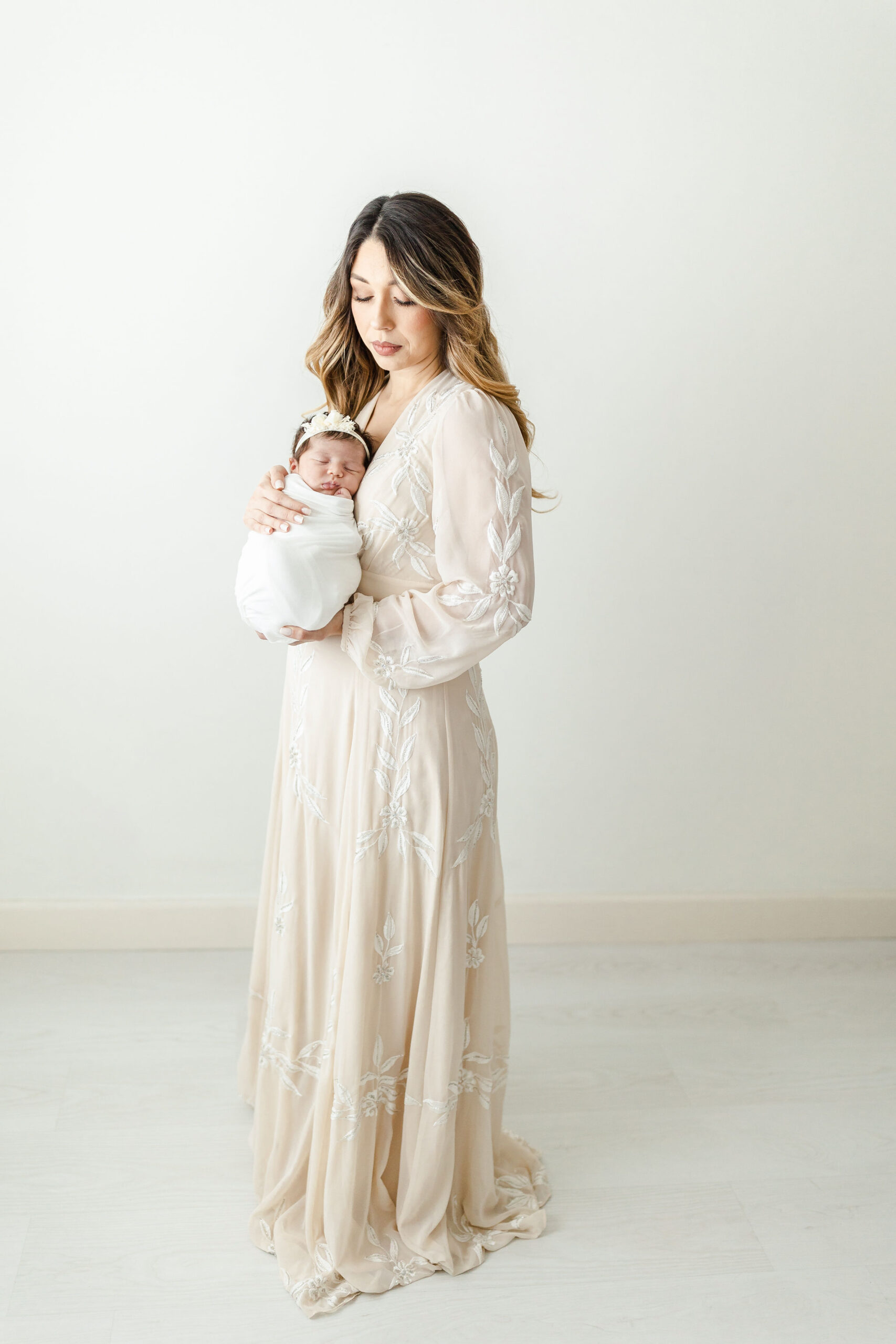 A new mom wearing a long cream down looks down at her sleeping newborn baby against her chest in a studio after getting placenta encapsulation miami
