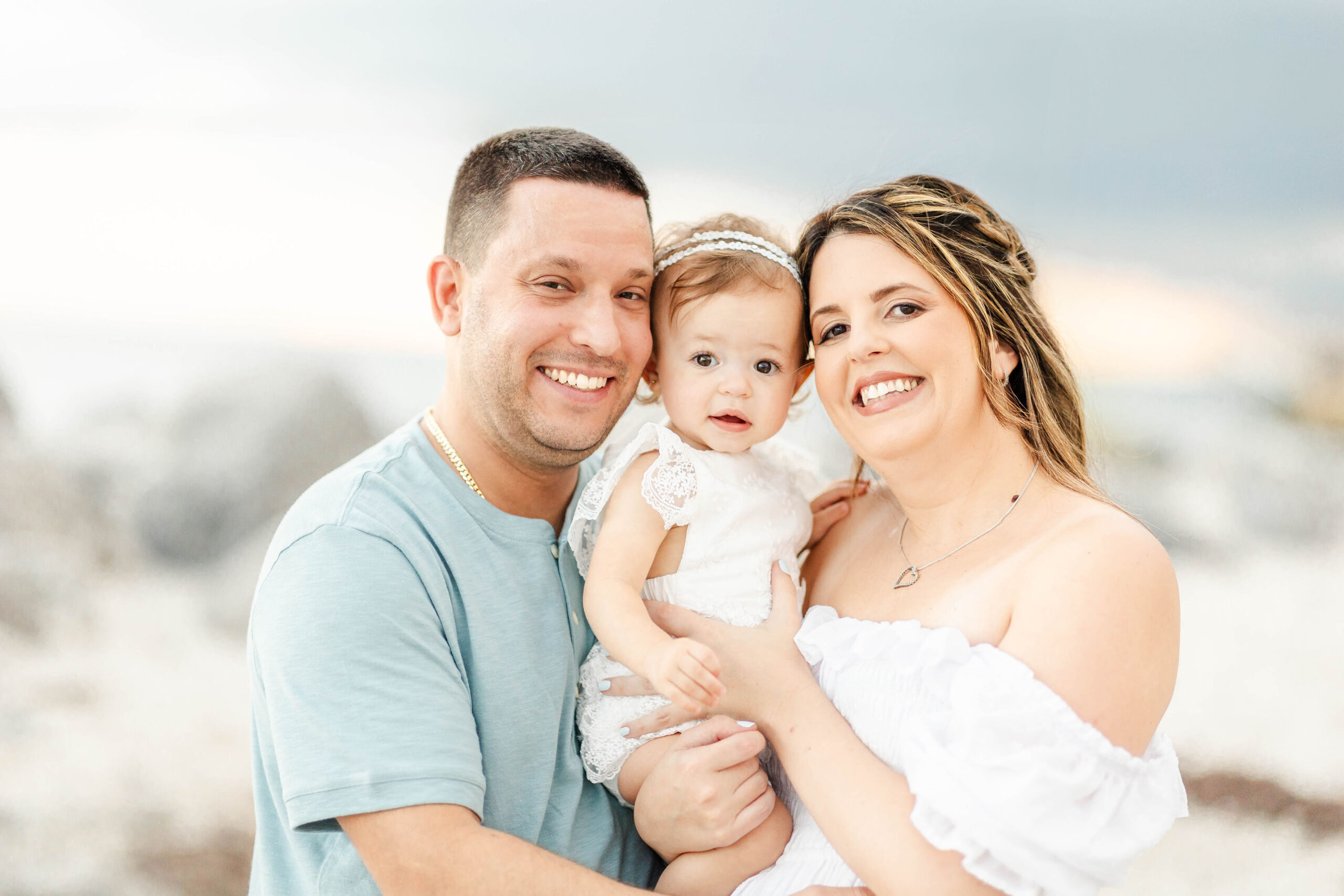 Smiling mom and dad stand on a beach holding their toddler daughter in a white dress after finding children's clothing miami