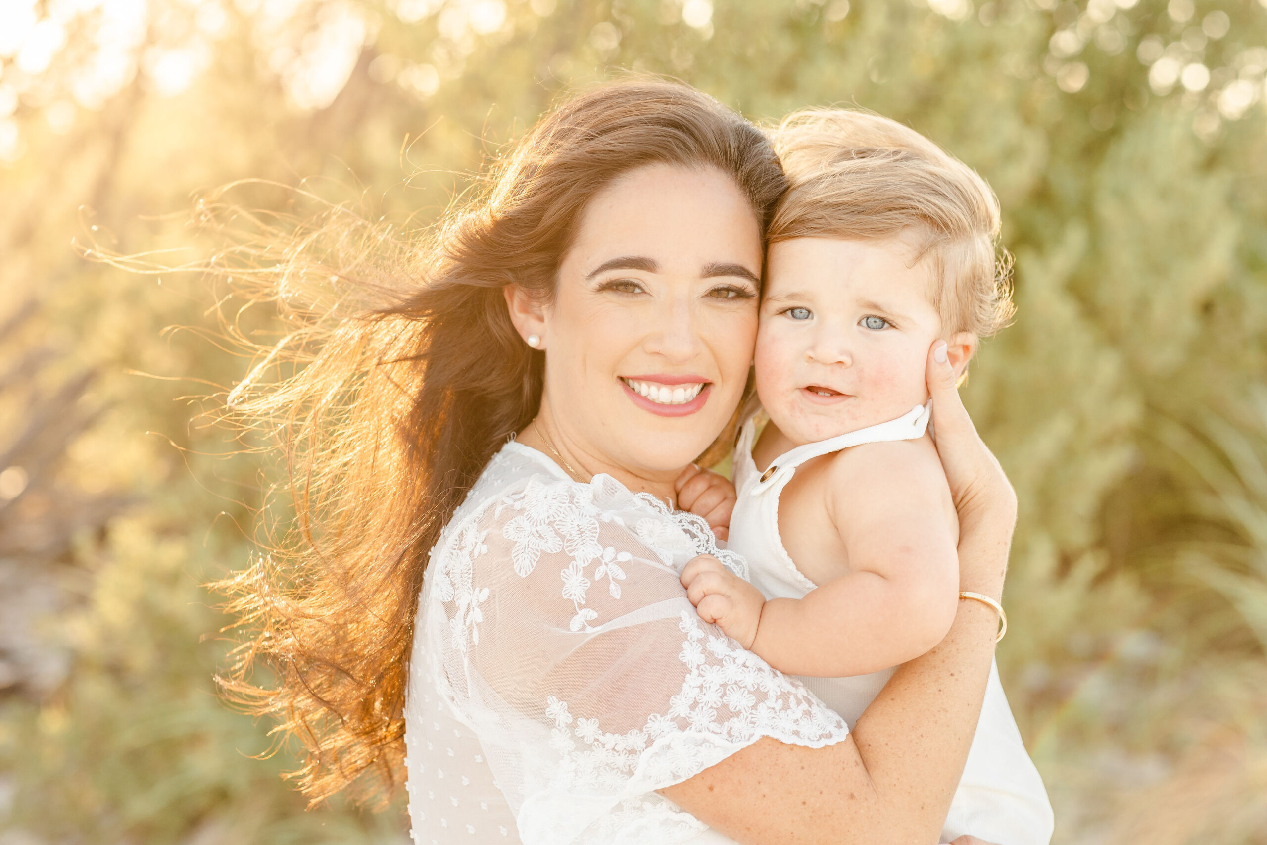 A happy mom smiles while snuggling with her toddler son on a windy beach at sunset while standing