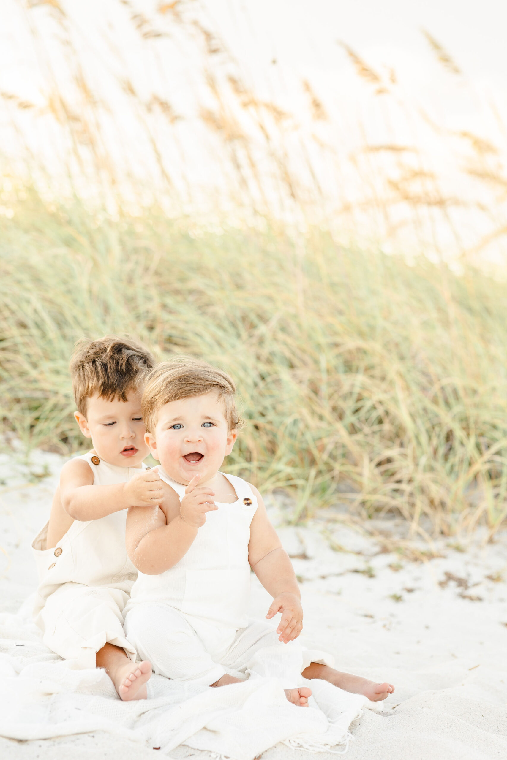 Happy toddler brothers in matching white overalls sit on a windy beach at sunset after visiting montessori schools in miami