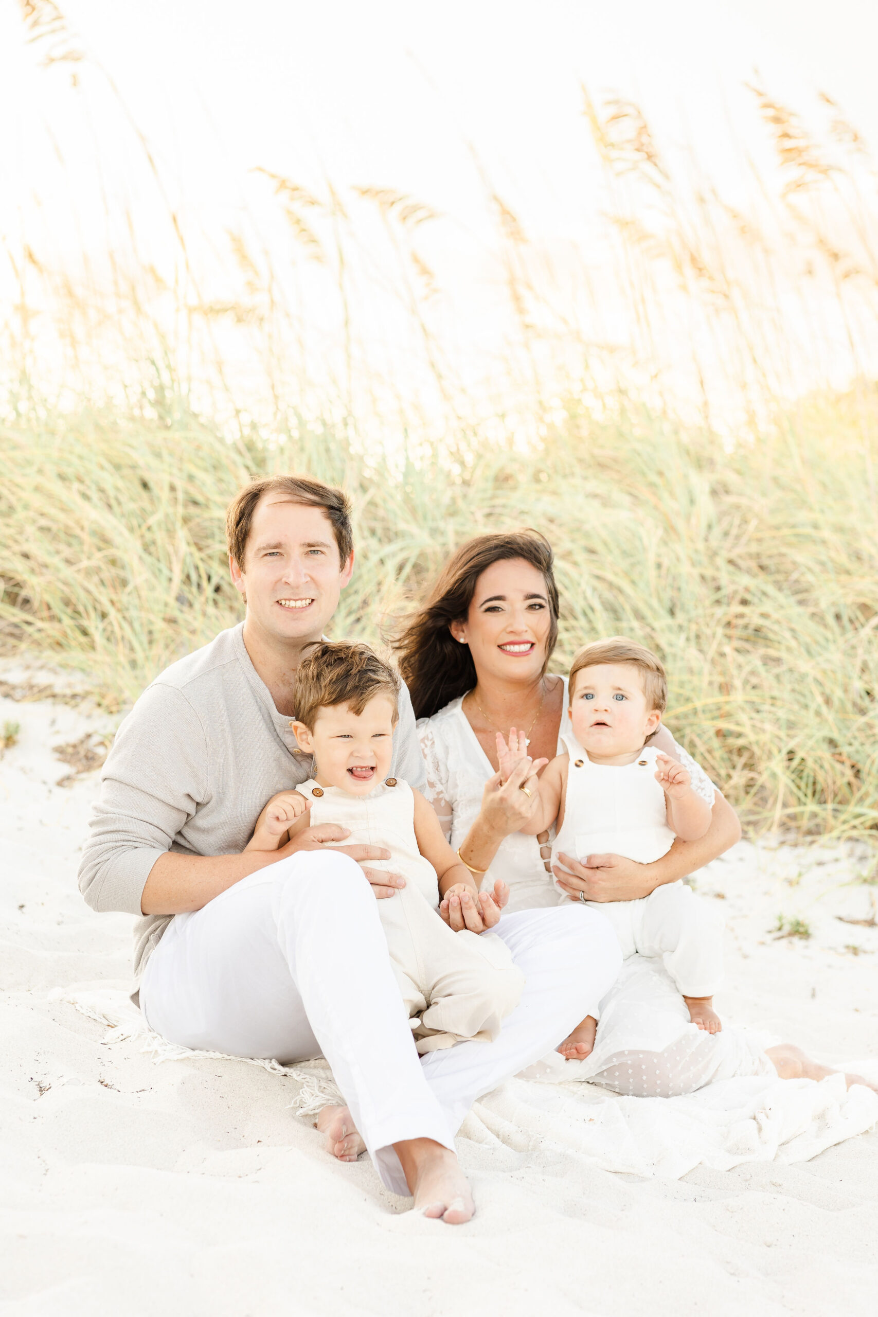 Happy parents sit on a windy beach in the sand with their toddler sons in their laps after visiting montessori schools in miami