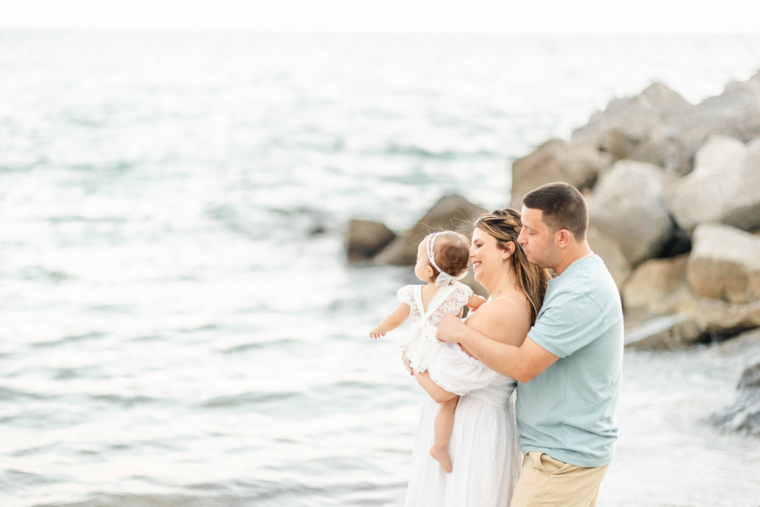 Happy mom and dad stand on a beach exploring with their toddler daughter on mom's hip before some swim lessons miami