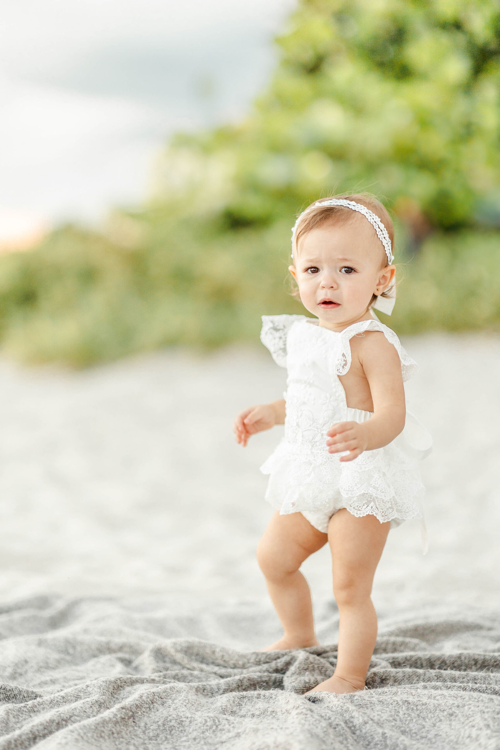 A toddler girl in a lace onesie walks on a blanket on the beach before some swim lessons miami