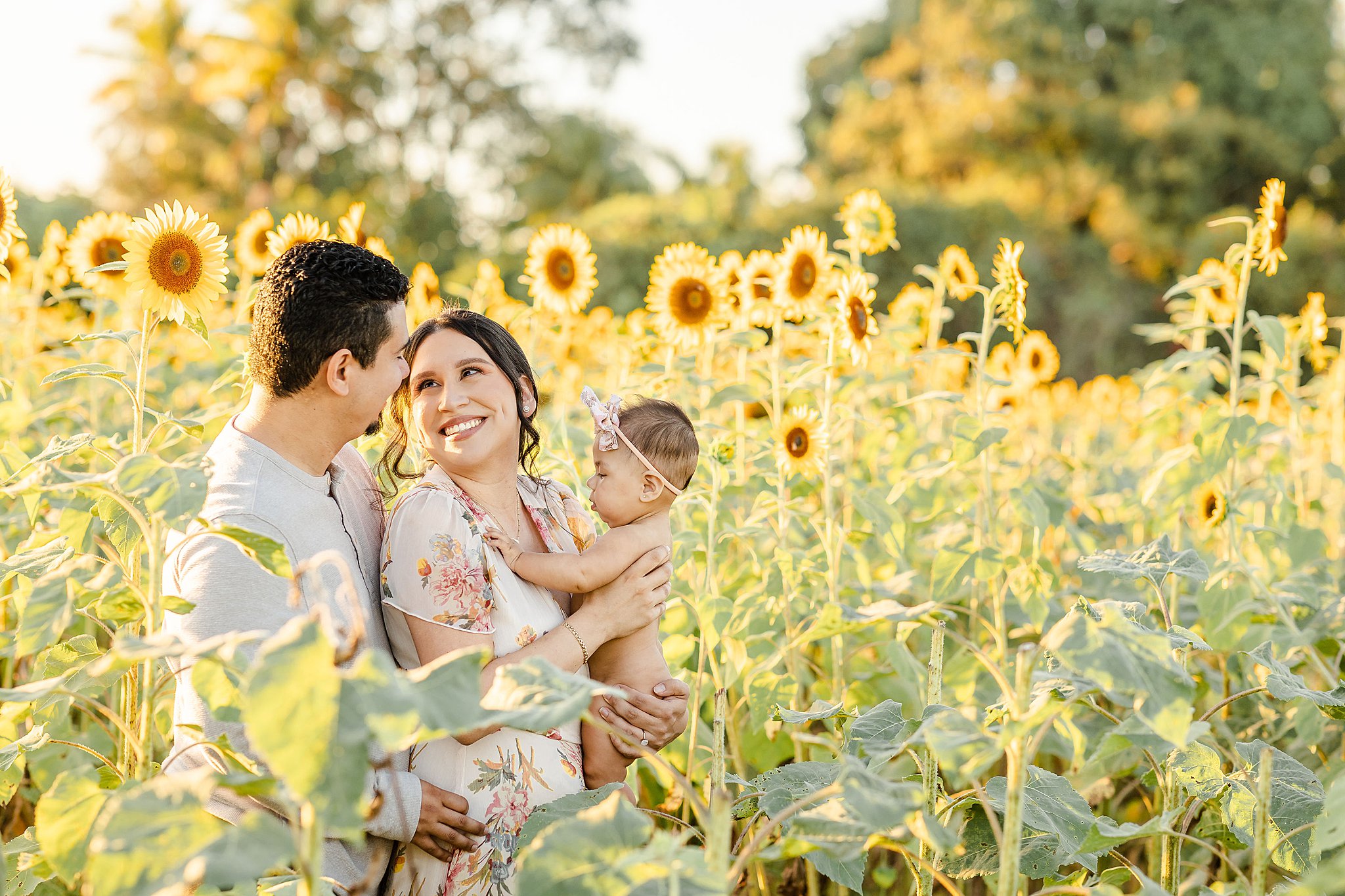 Happy mom and dad snuggle in a sunflower field with their infant daughter in mom's arms during fall activities miami