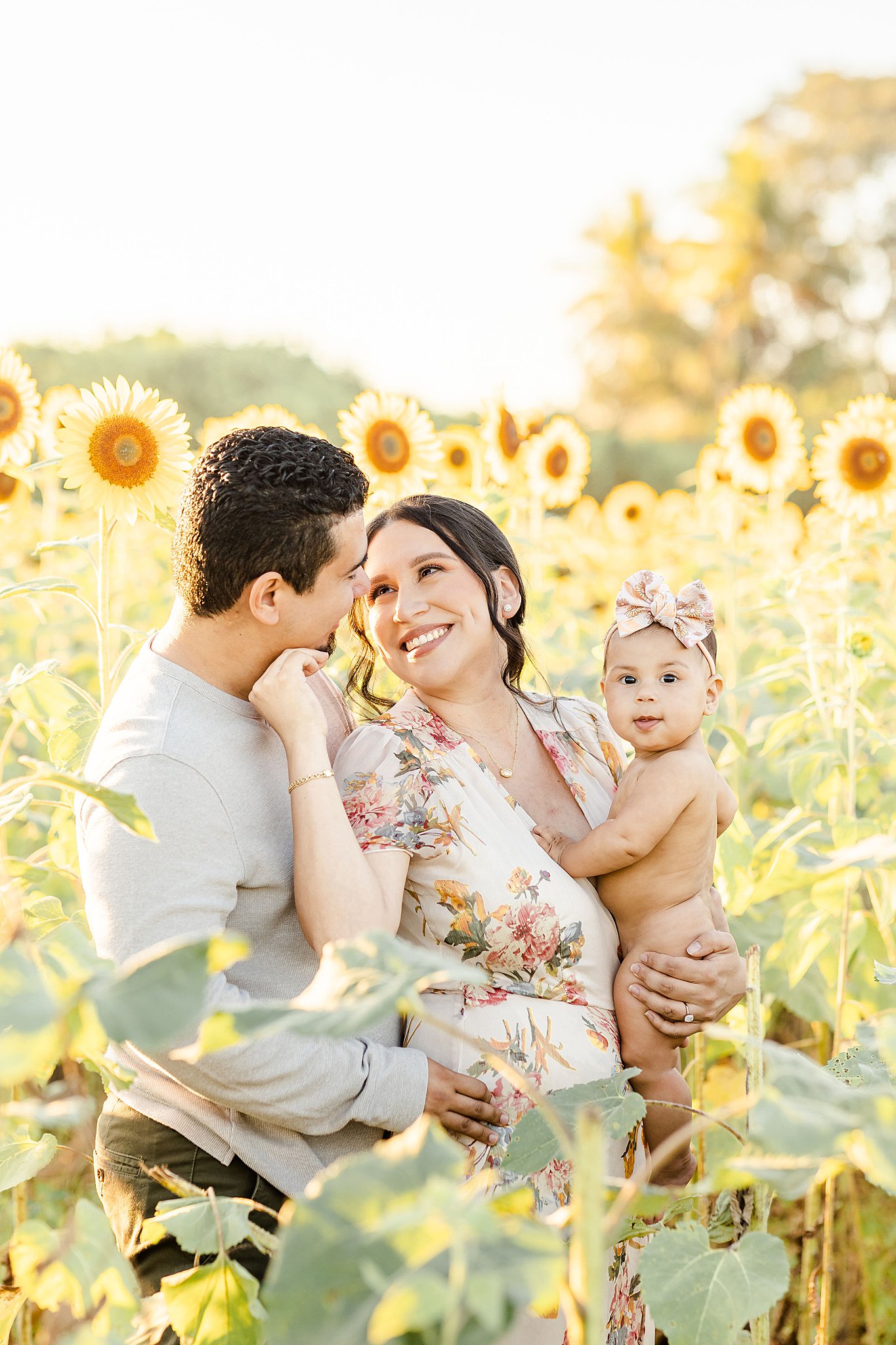 A naked infant girl sits in mom's arms while she leans into dad in a sunflower field at sunset during fall activities miami