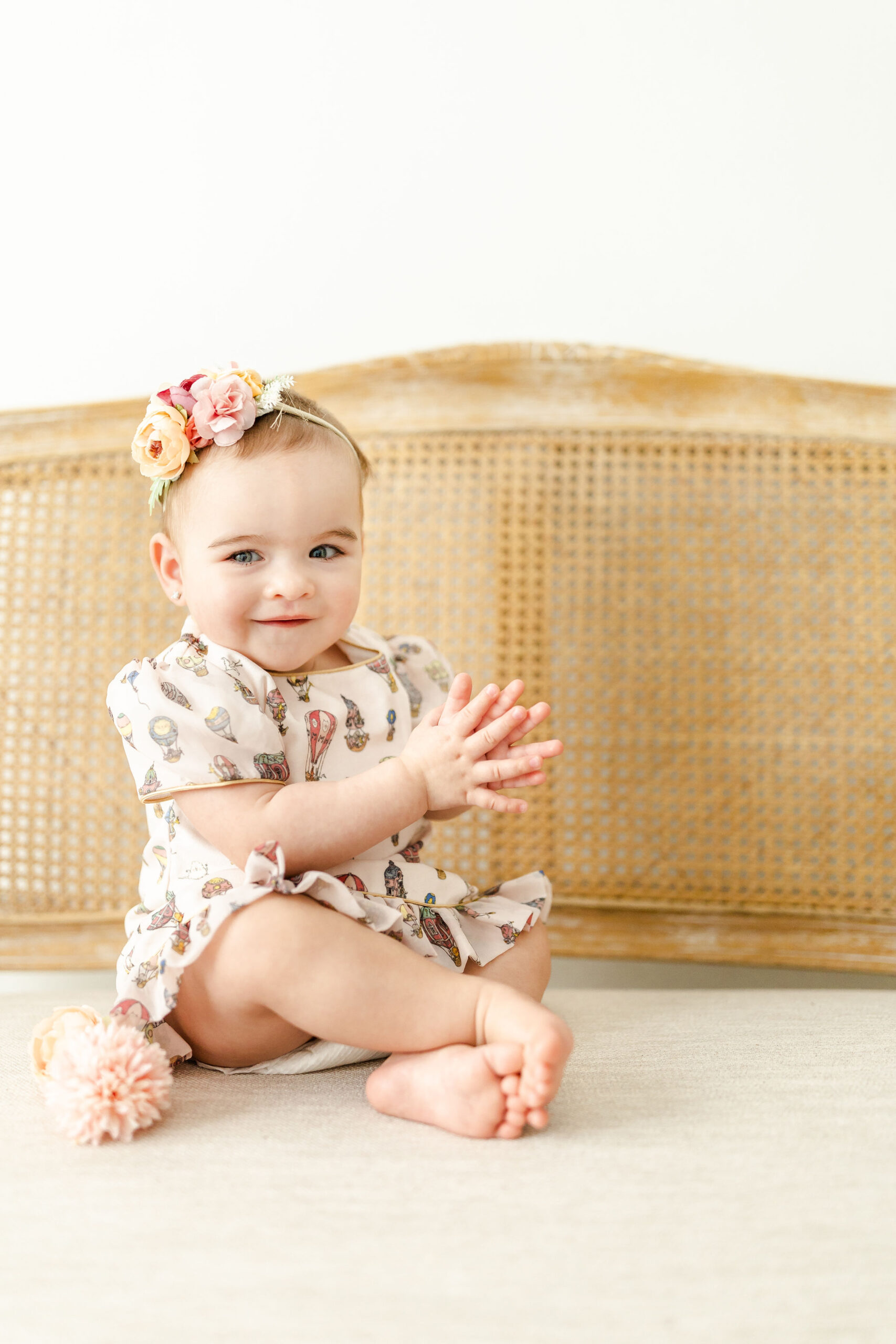 A happy toddler girl in a print dress sits on a bench in a studio clapping