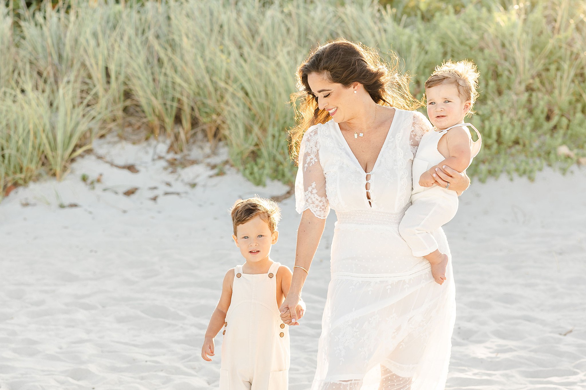 A smiling mom walks on a beach in a white dress at sunset with her 2 toddler sons