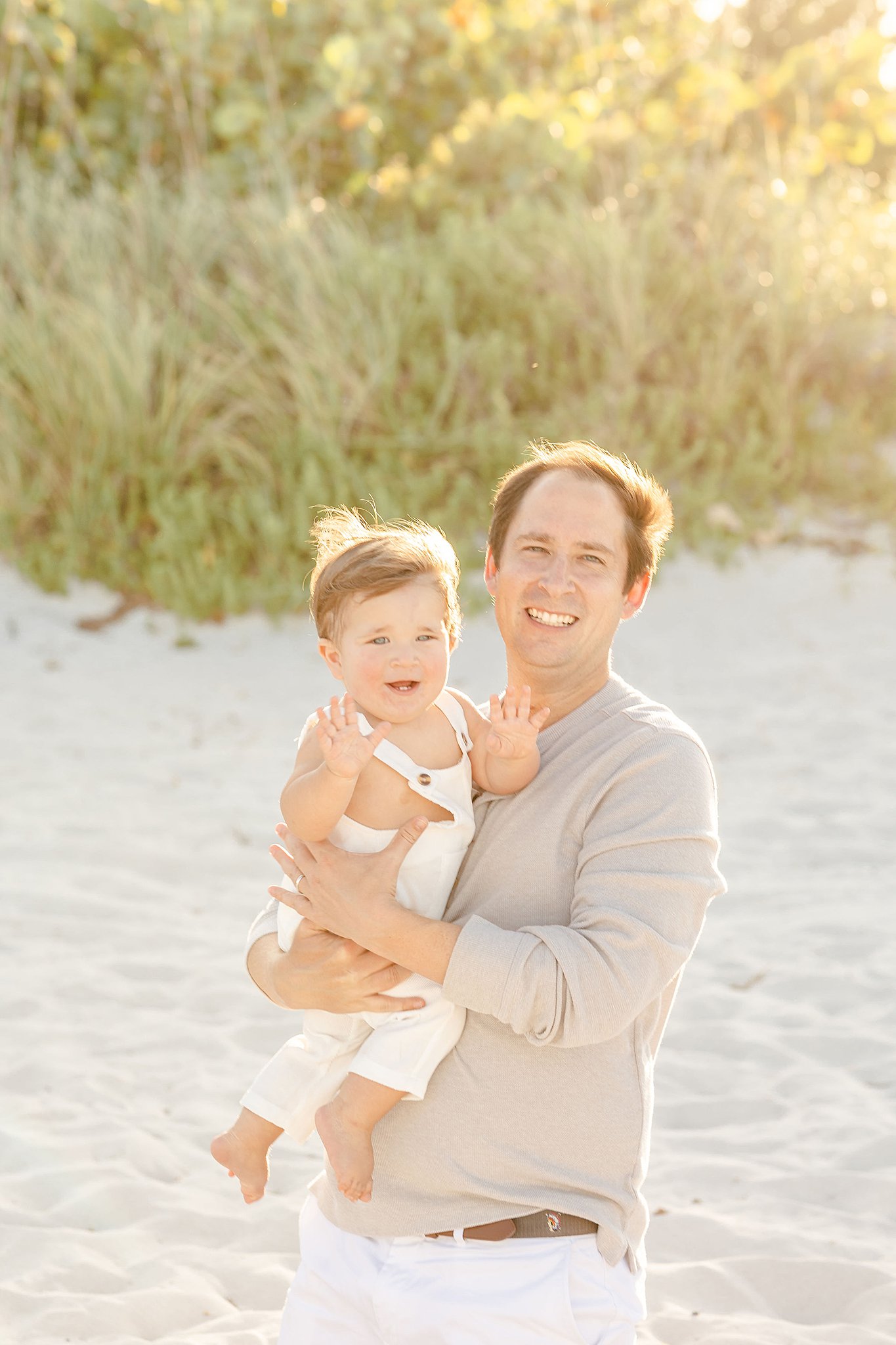 A toddler boy in white overalls plays with dad on a windy beach at sunset while staying at kid friendly hotels miami